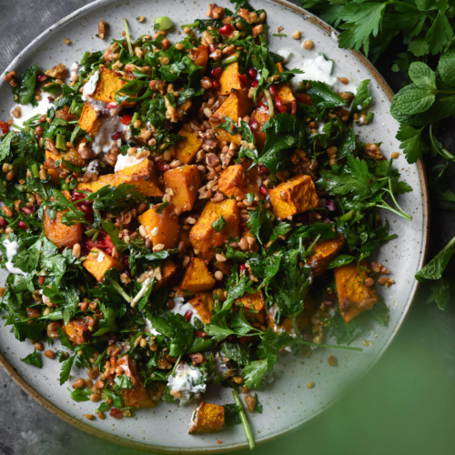 Preserved lemon roasted pumpkin with herb and toasted nut salad, yoghurt and pomegranate arils on a blue backdrop surrounded by herbs