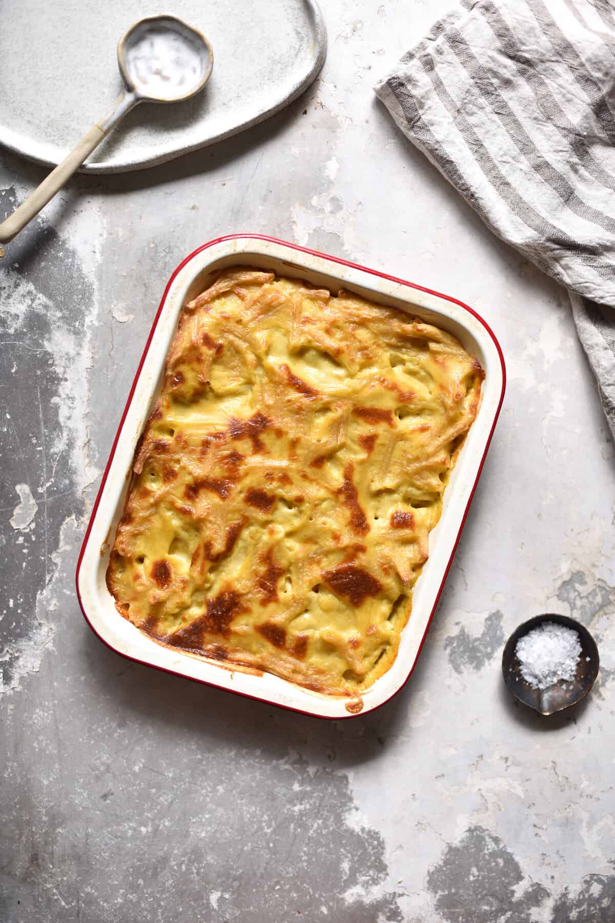 Gluten free vegan mac and cheese in a rectangular baking dish on a mottled white and silver backdrop. A linen cloth, small salt plates and an empty ceramic plate surround the vegan mac and cheese