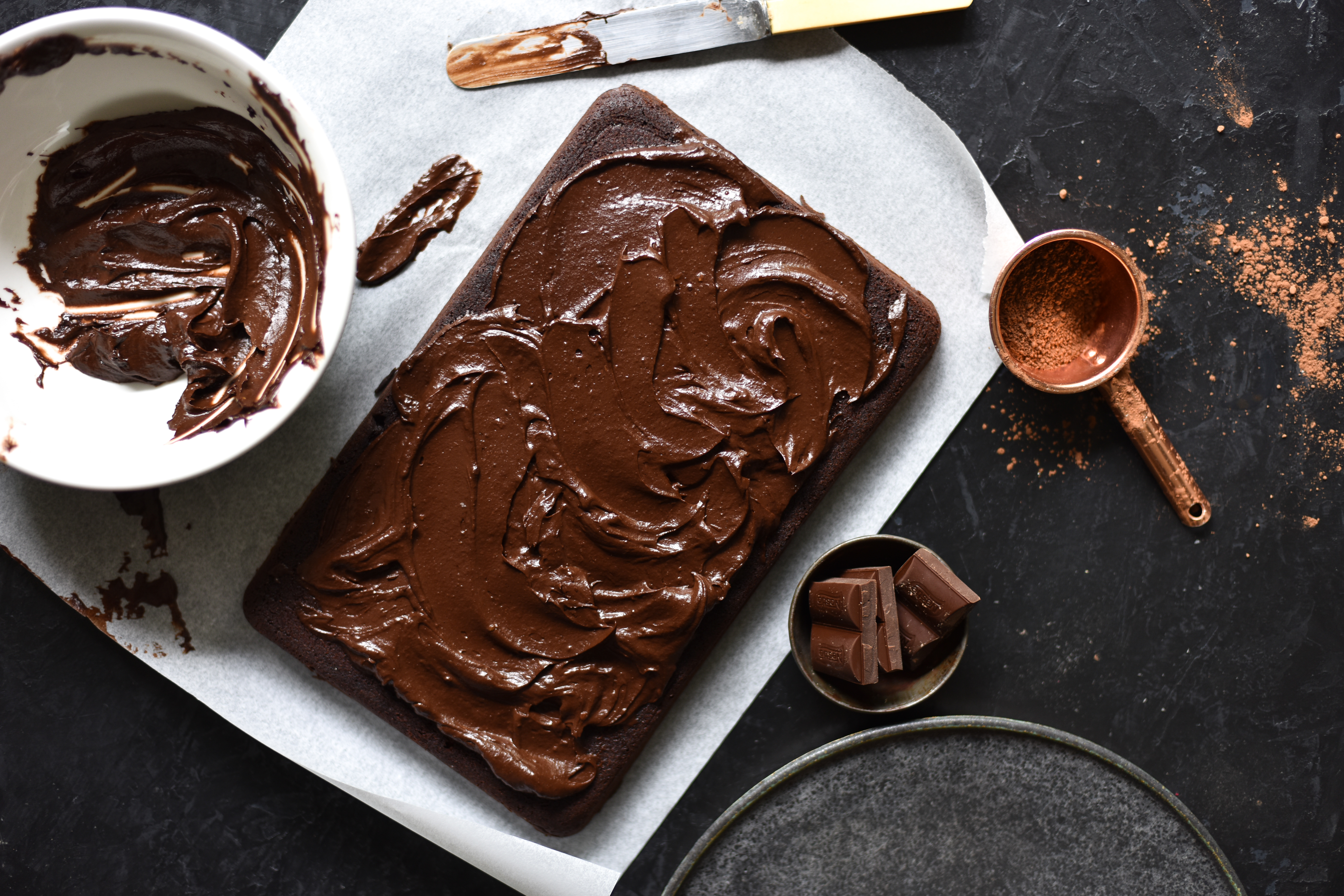 An aerial image of a gluten free zucchini chocolate cake with chocolate almond icing on a dark backdrop surrounded by baking ingredients. 