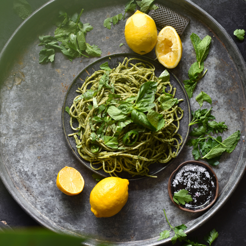Gluten free and vegan avocado and herb sauced pasta on a blue backdrop, surrounded by lemons, herbs and greenery