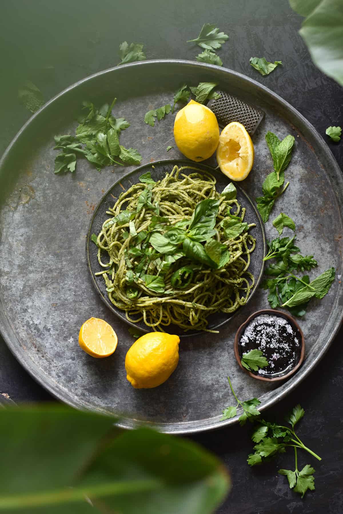 Gluten free and vegan avocado and herb sauced pasta on a blue backdrop, surrounded by lemons, herbs and greenery