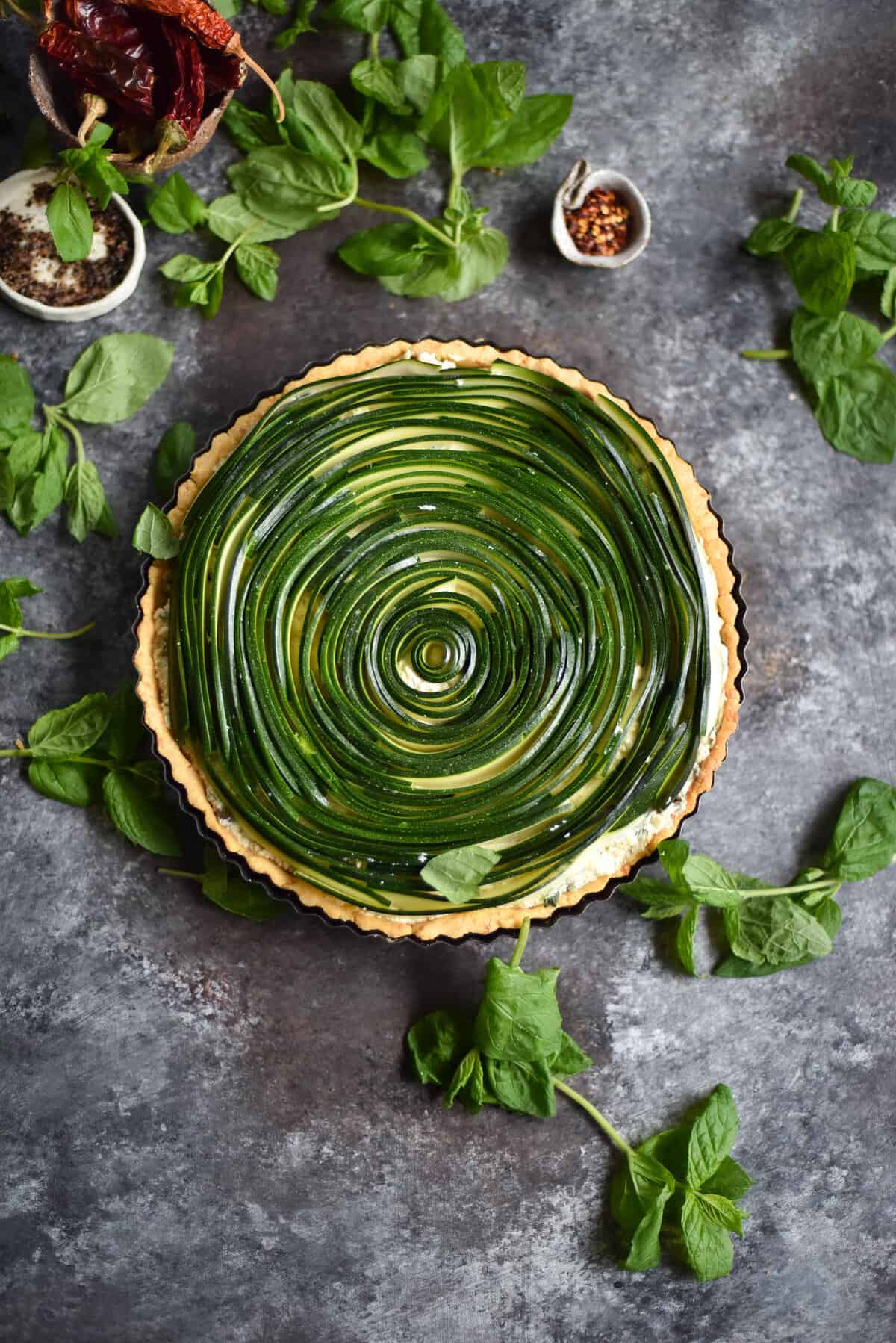 Gluten free spiral zucchini tart with ricotta, herbs and capers on a blue backdrop and scattered with herbs