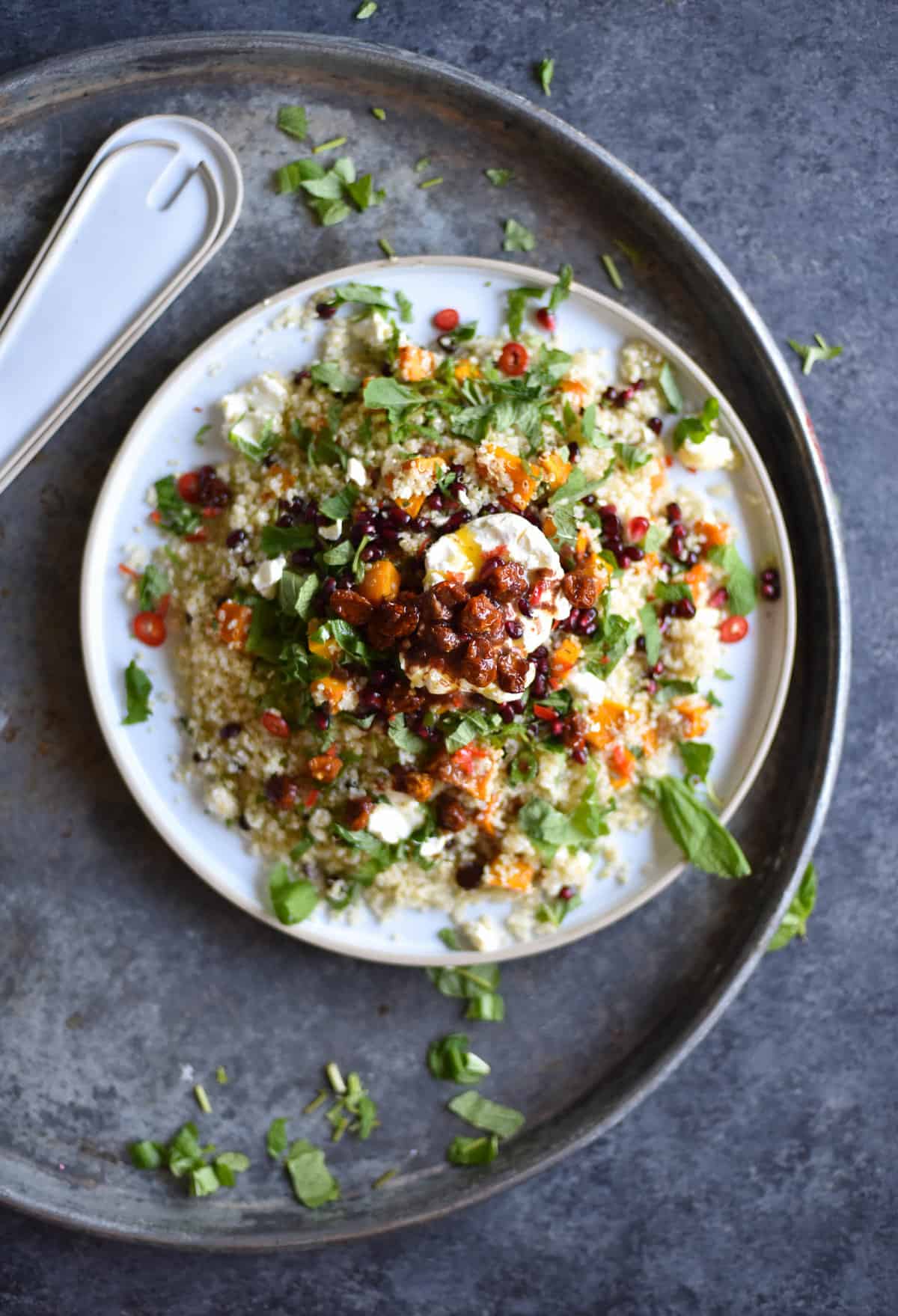 An aerial image of a quinoa salad with roasted vegetables and preserved lemon butter on a white ceramic plate atop a dark blue backdrop