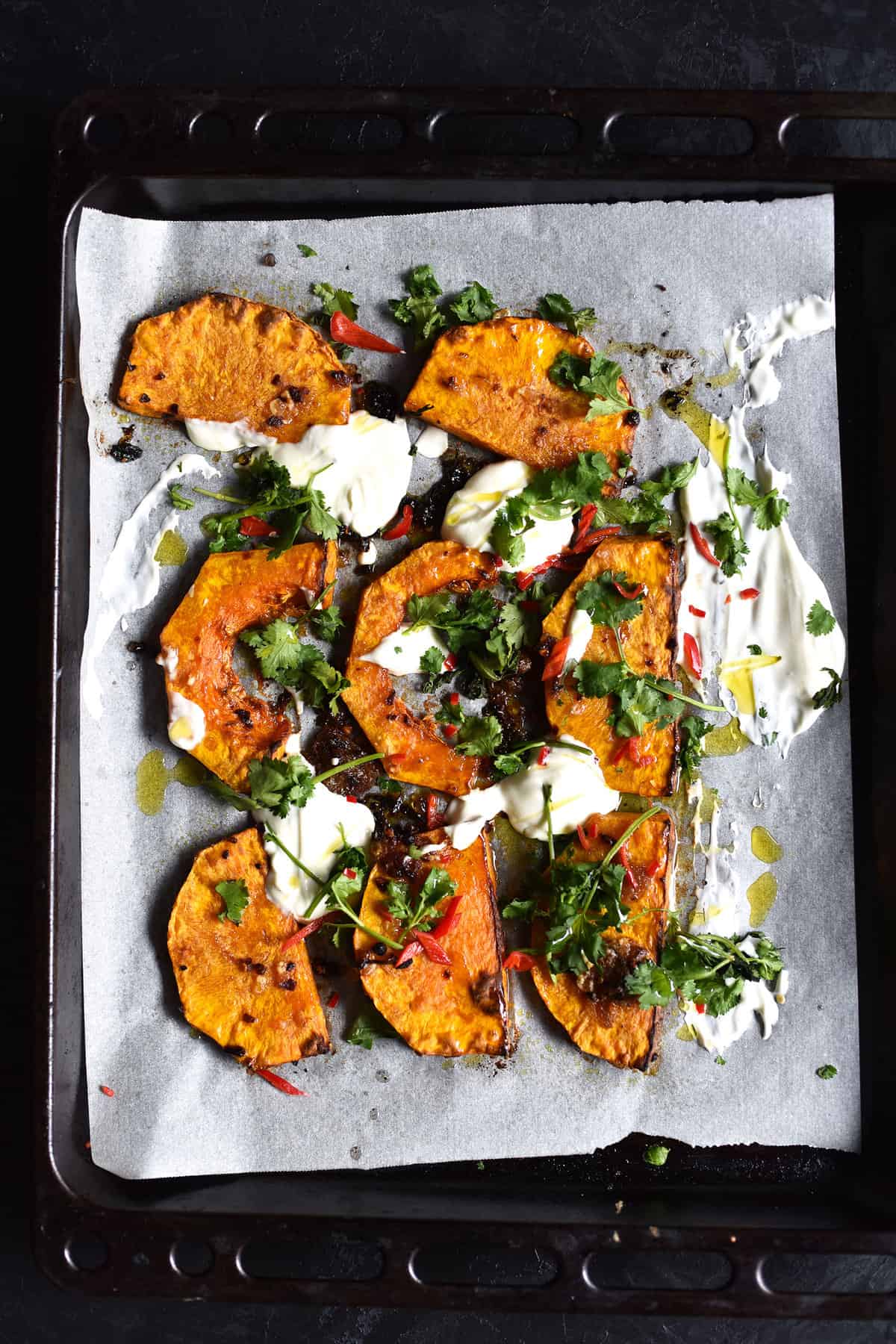 An aerial image of preserved lemon roasted pumpkin with Greek yoghurt, herbs and chilli on a black baking tray atop a black backdrop