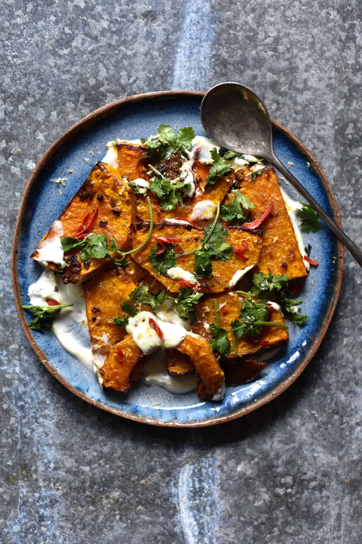 An aerial view of a blue ceramic plate topped with preserved lemon roasted pumpkin, Greek yoghurt, chilli and chopped herbs. The plate sits on a speckled grey steel backdrop