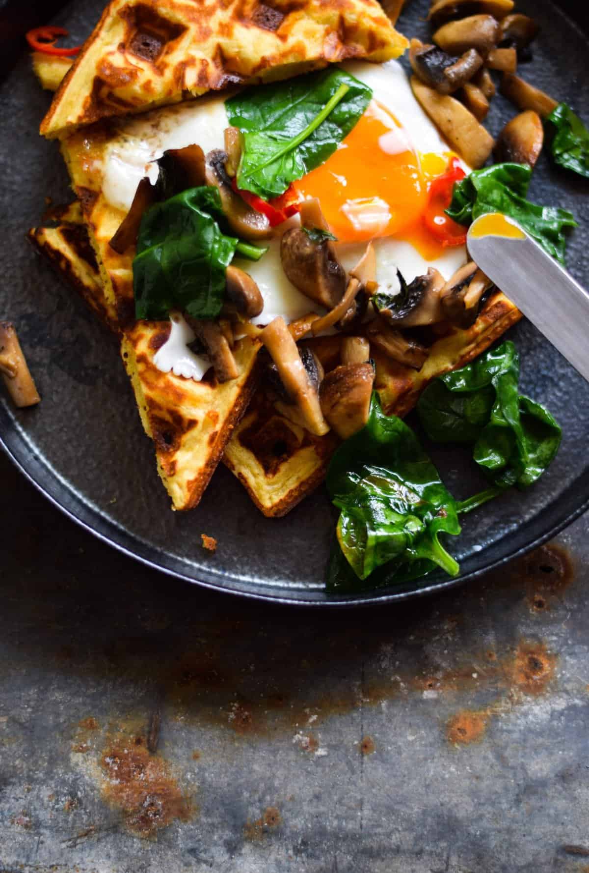 An aerial image of a dark ceramic plate topped with chickpea flour waffles. The waffles are served with a runny fried egg, greens and some mushrooms. They sit atop a dark blue table