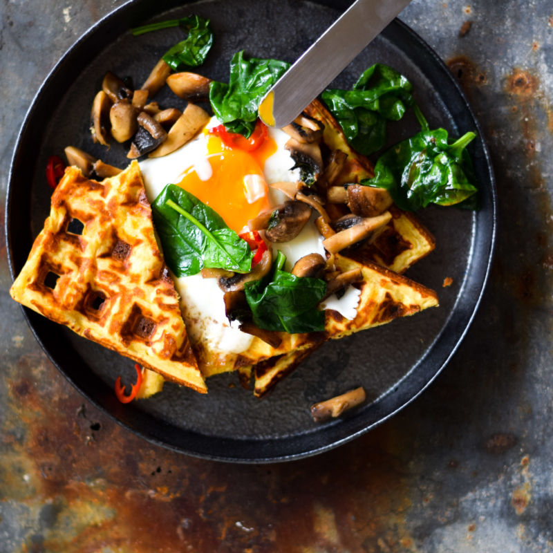 An aerial image of a dark ceramic plate topped with chickpea flour waffles. The waffles are served with a runny fried egg, greens and some mushrooms. They sit atop a dark blue table