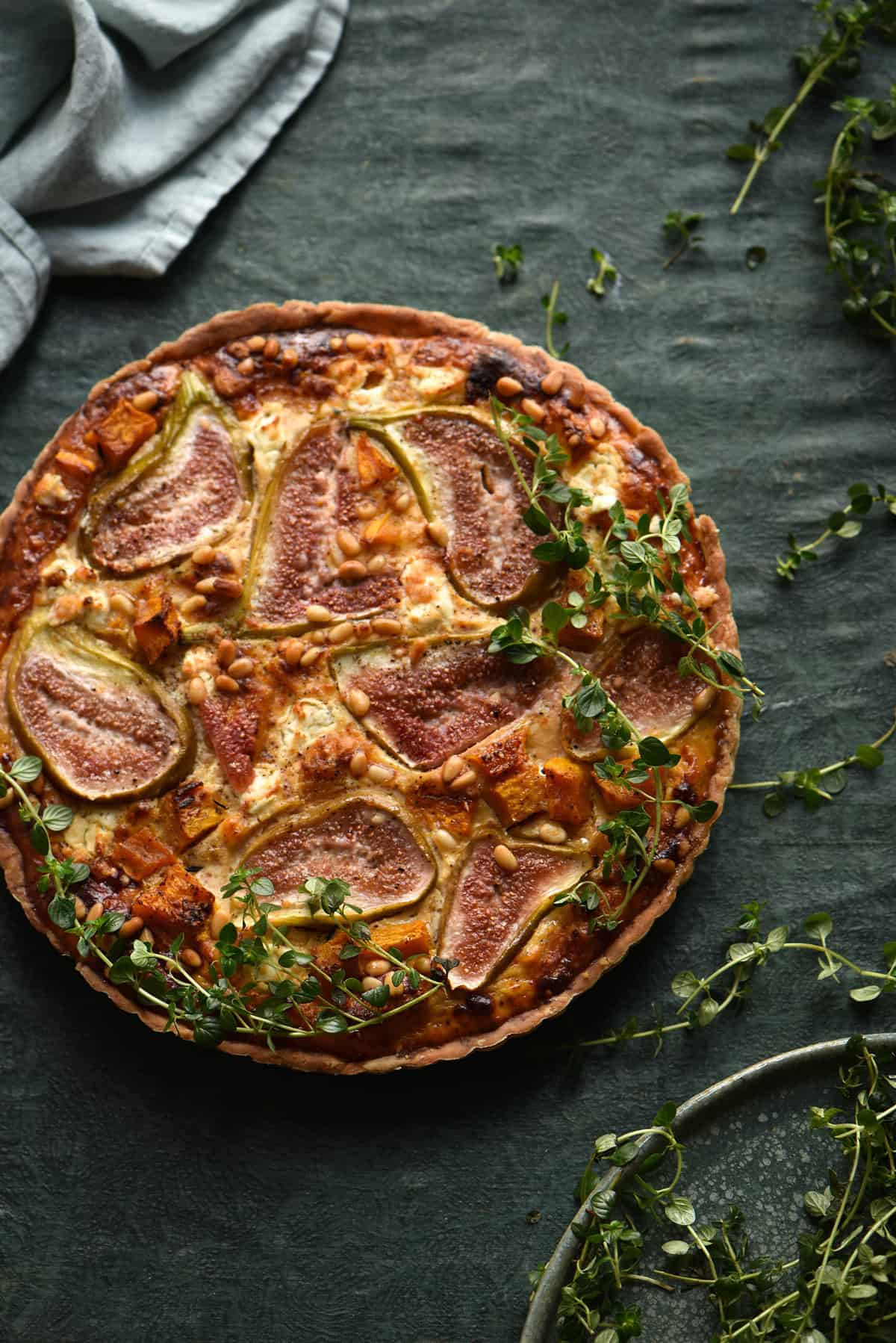 Savoury fig, pumpkin and goats cheese tart in a gluten free crust on a green backdrop with scattered herbs
