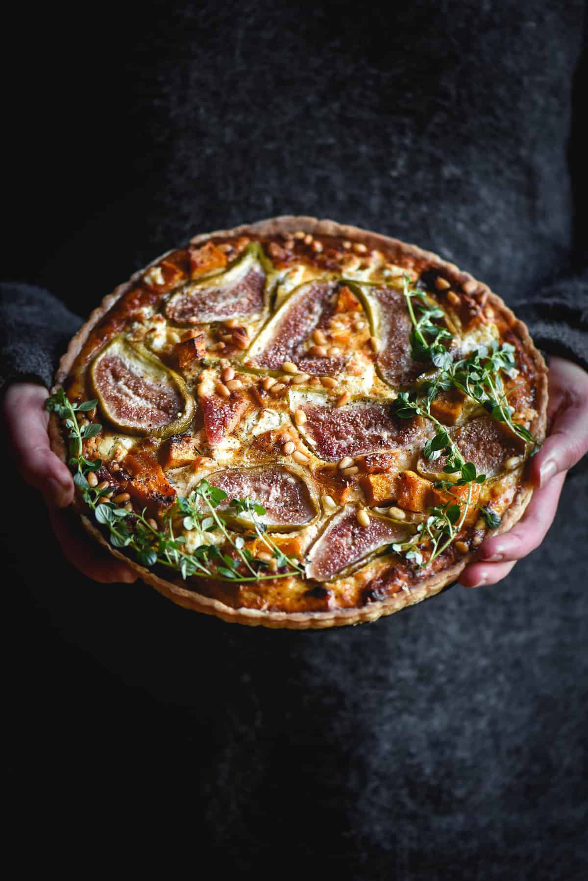 Gluten free fig, pumpkin, pine nut and goats cheese tart being held by a woman in a dark fluffy jumper