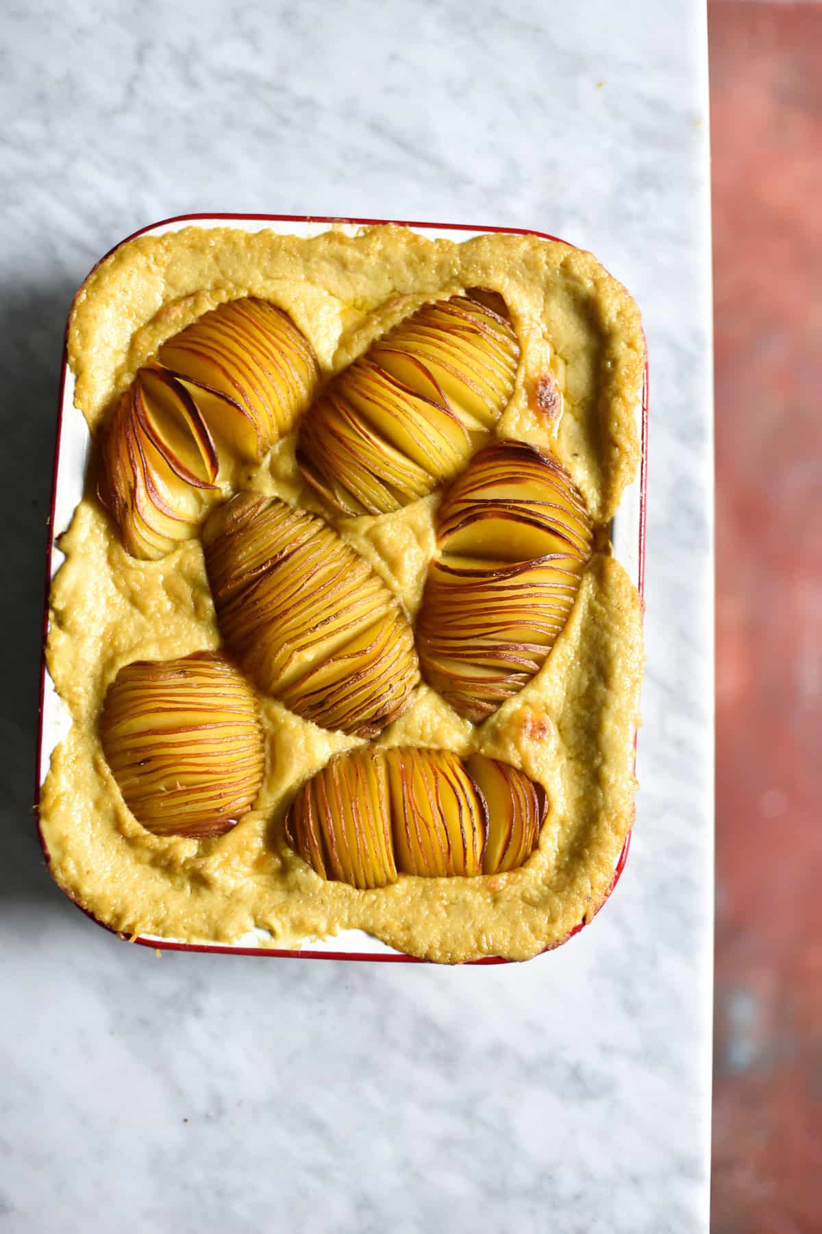 Gluten free, vegan, FODMAP friendly cheesy hasselback potato bake in a white baking dish atop a white marble table