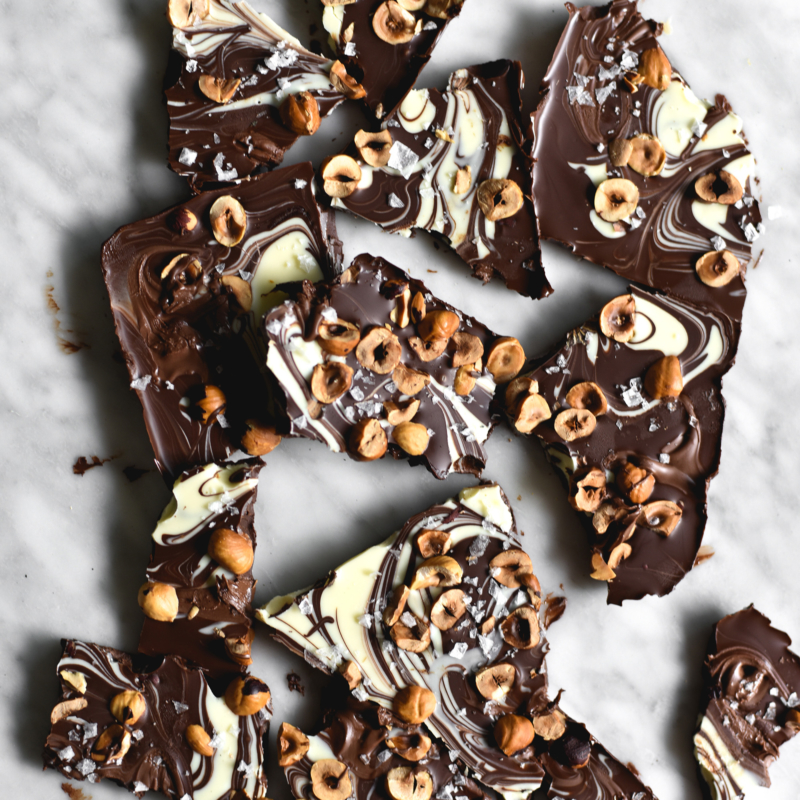 An aerial image of marbled chocolate bark topped with toasted hazelnuts that has been snapped into pieces and arranged on a white marble table.