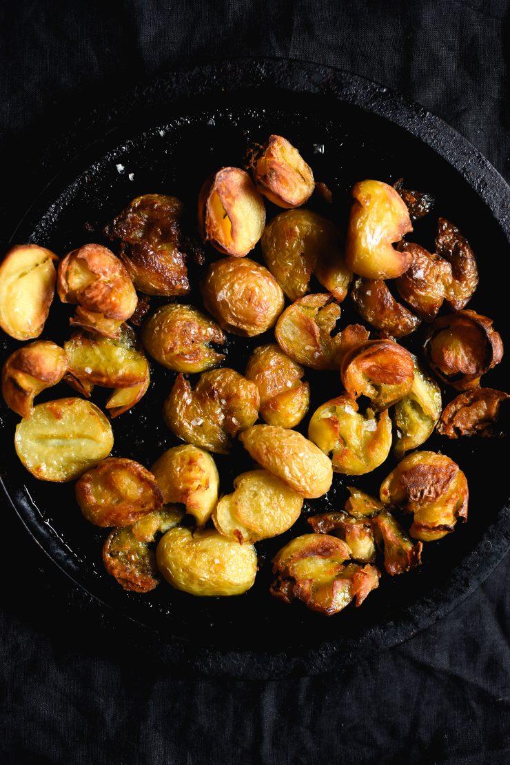 Crispy salt and vinegar roasted potatoes on a black round baking tray sitting a black backdrop