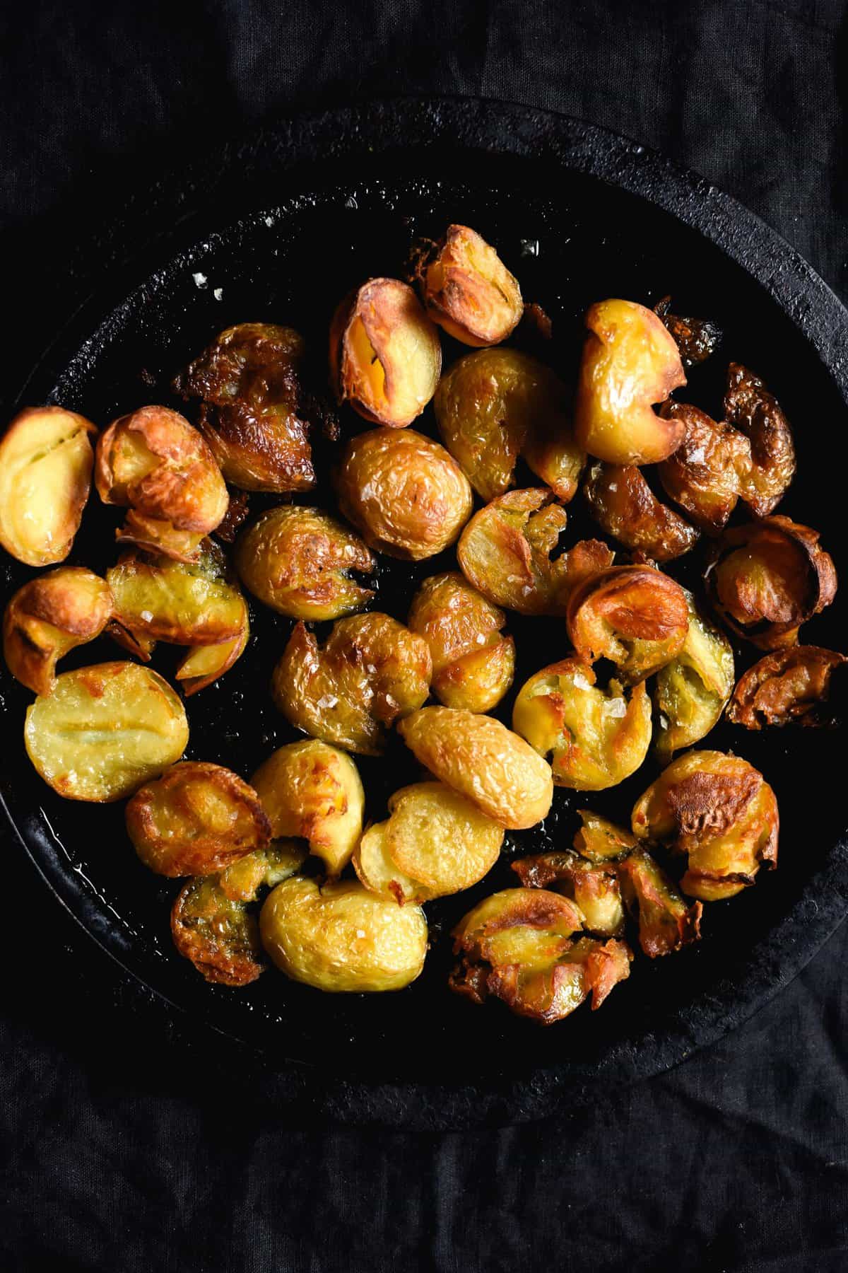 Crispy salt and vinegar roasted potatoes on a black round baking tray sitting on a black backdrop