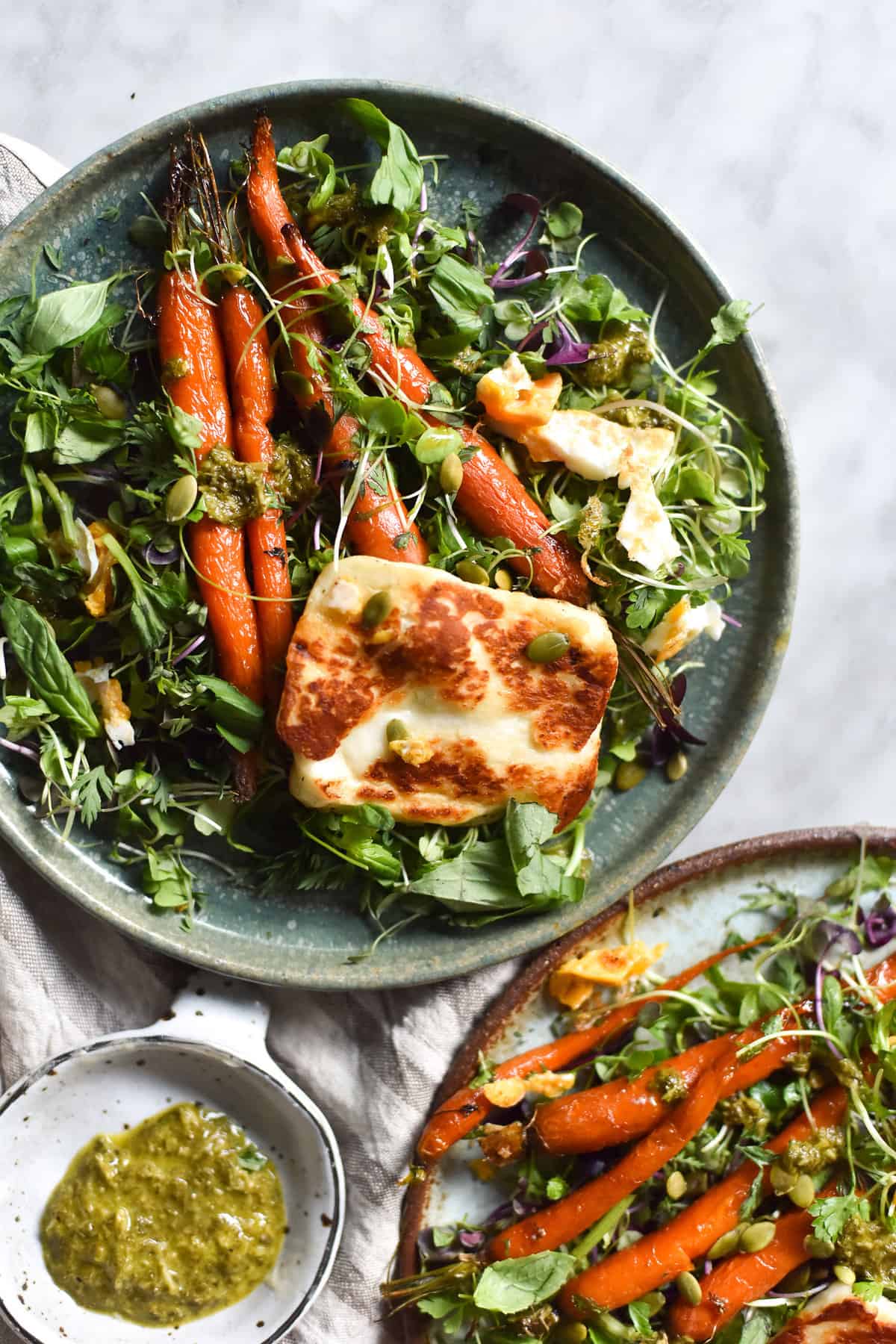 An aerial image of two plates of honey butter roasted carrot salad with shaved fennel, haloumi and a herby preserved lemon sauce. The plates sit atop a white marble table. 