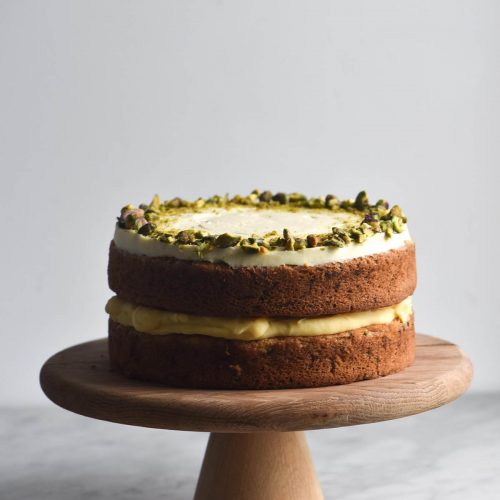 A gluten free version of Nigella's courgette cake on a wooden cake stand on a marble table, against a white backdrop. The cake has two layers, separated in the middle by dairy free lemon curd, and topped with cream cheese icing and a crown of chopped pistachios