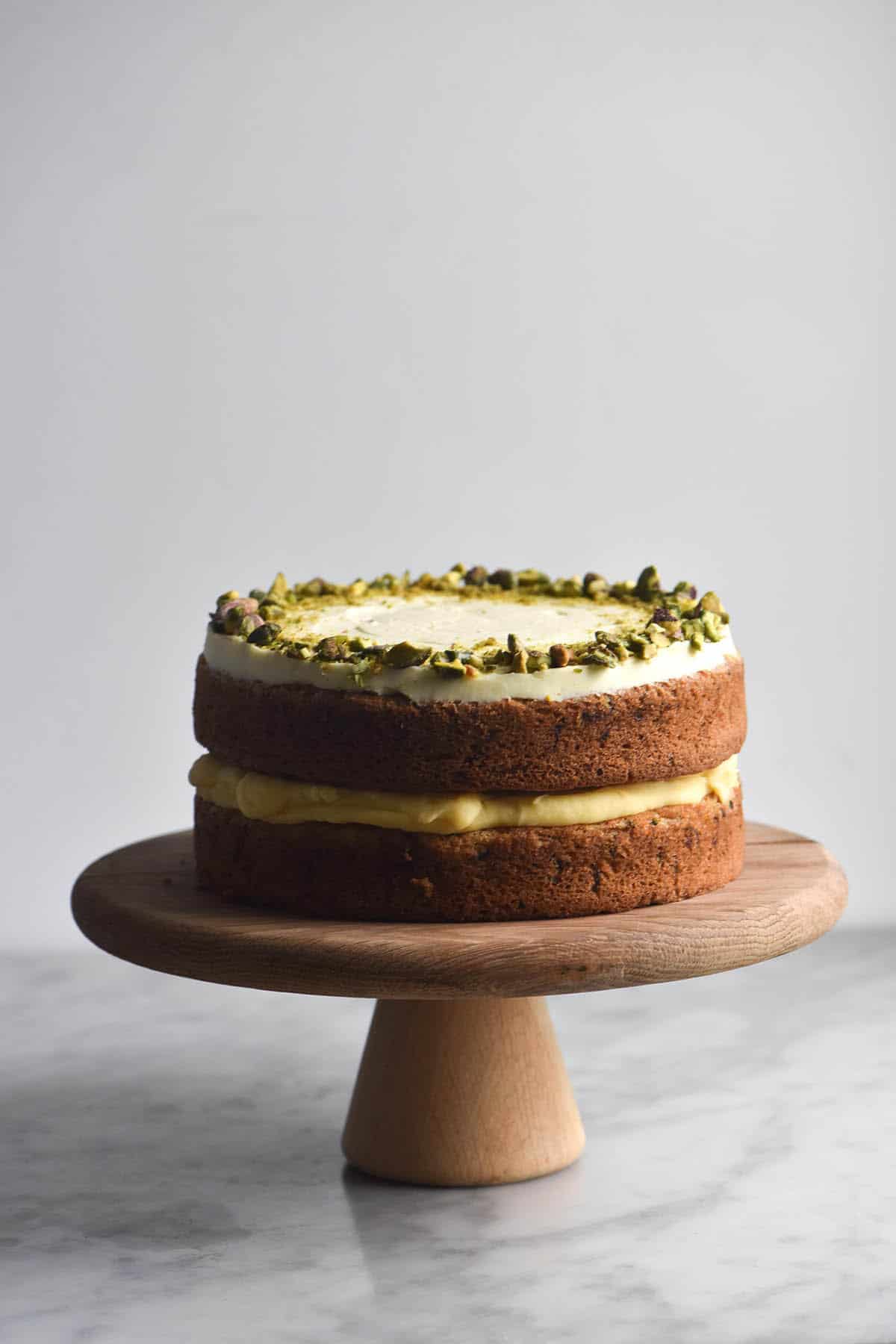 A gluten free version of Nigella's courgette cake on a wooden cake stand on a marble table, against a white backdrop. The cake has two layers, separated in the middle by dairy free lemon curd, and topped with cream cheese icing and a crown of chopped pistachios