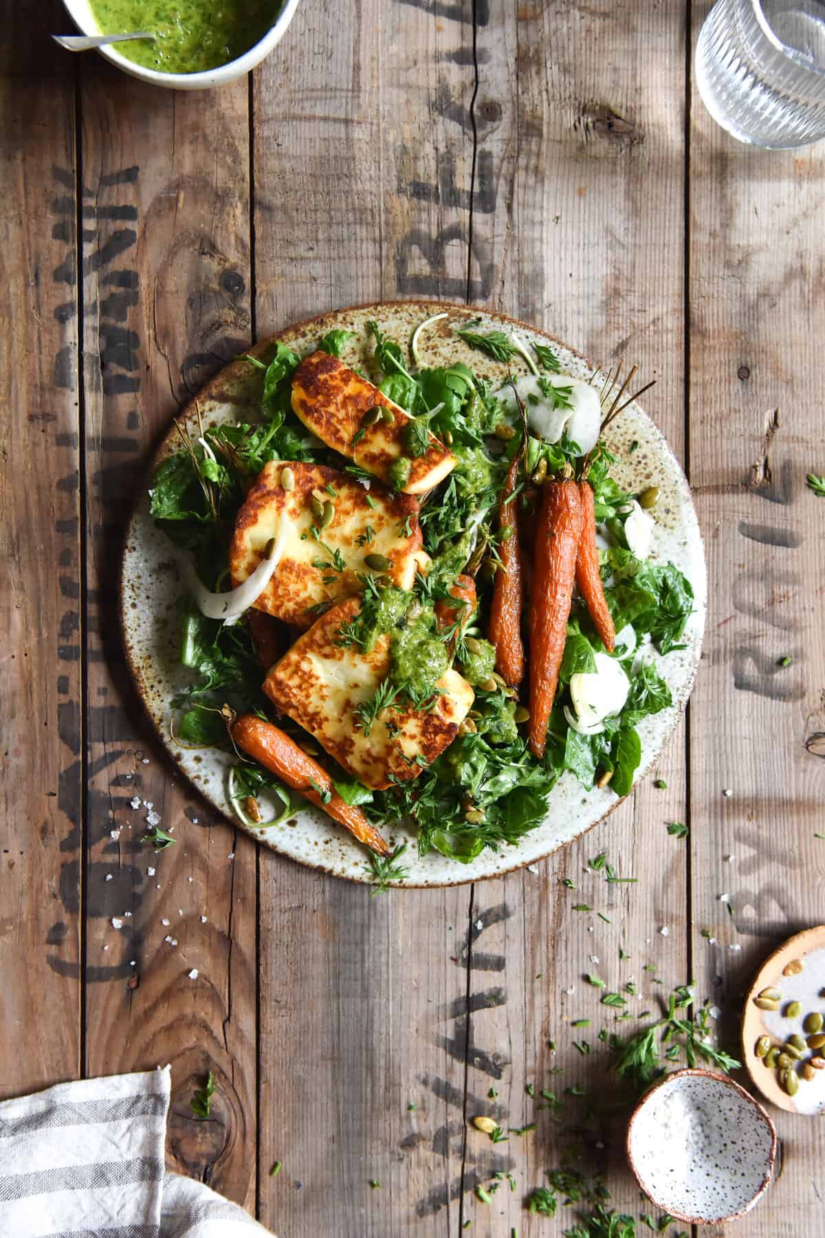 An aerial image of a roasted carrot salad with haloumi and green herb sauce on a wooden table. The salad sits in the centre of the image and is surrounded by scattered herbs, small pinch bowls and a beige linen tea towel.