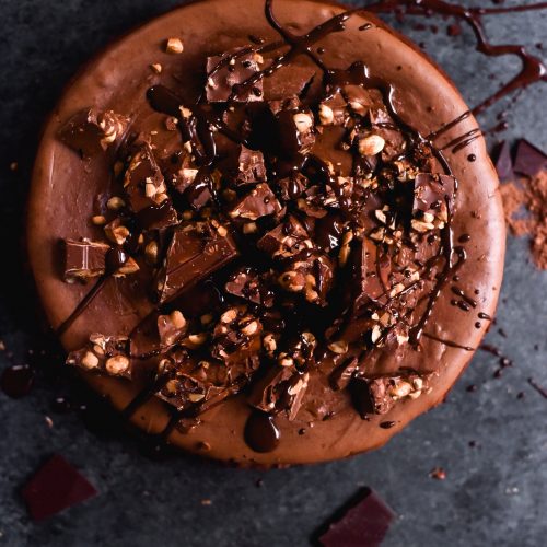 An aerial image of a gluten free chocolate and peanut butter cheesecake on a dark blue backdrop. The cheesecake is topped with Whittakers bars and melted chocolate drizzles.