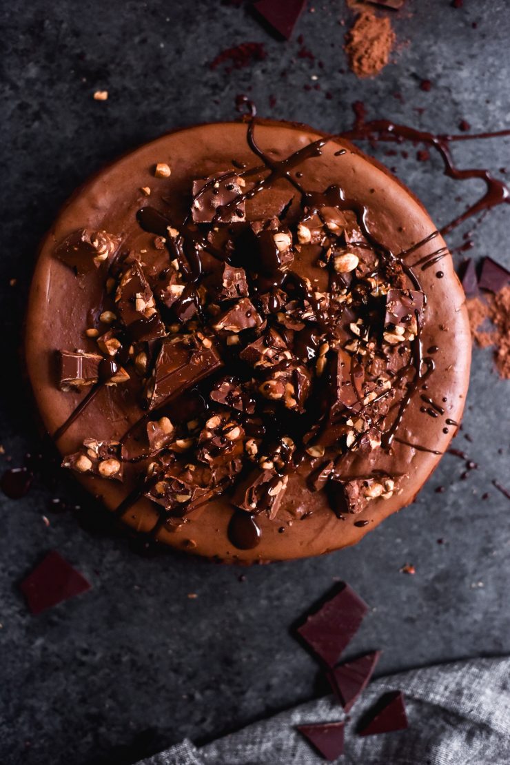 An aerial image of a gluten free chocolate and peanut butter cheesecake on a dark blue backdrop. The cheesecake is topped with Whittakers bars and melted chocolate drizzles.
