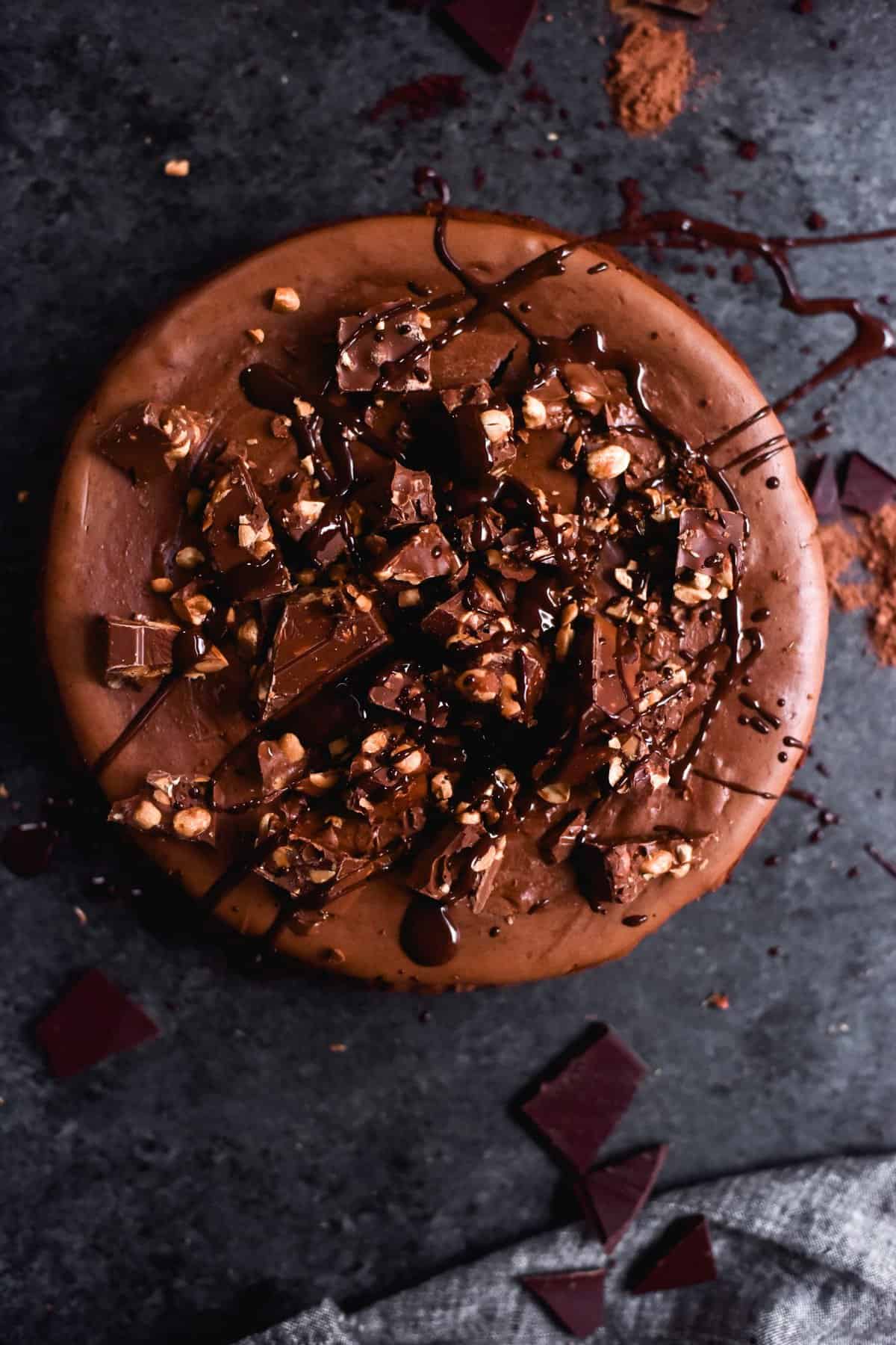 An aerial image of a gluten free chocolate and peanut butter cheesecake on a dark blue backdrop. The cheesecake is topped with Whittakers bars and melted chocolate drizzles.