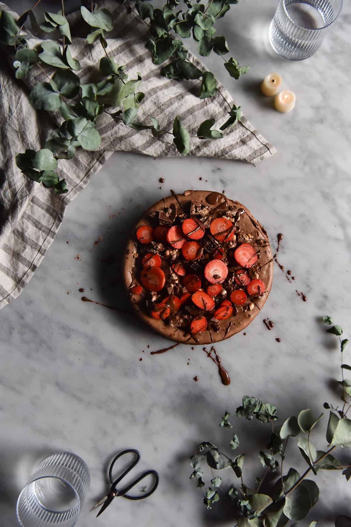 An aerial image of a gluten free chocolate peanut butter cheesecake on a marble table. The cheesecake is topped with strawberries and drizzles of melted chocolate.