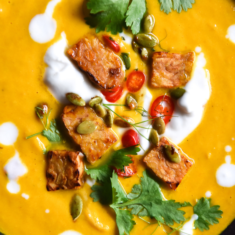 A close up aerial photo of a bowl of vegan Thai carrot soup with crunchy tempeh bits, cream, herbs and chopped chilli.