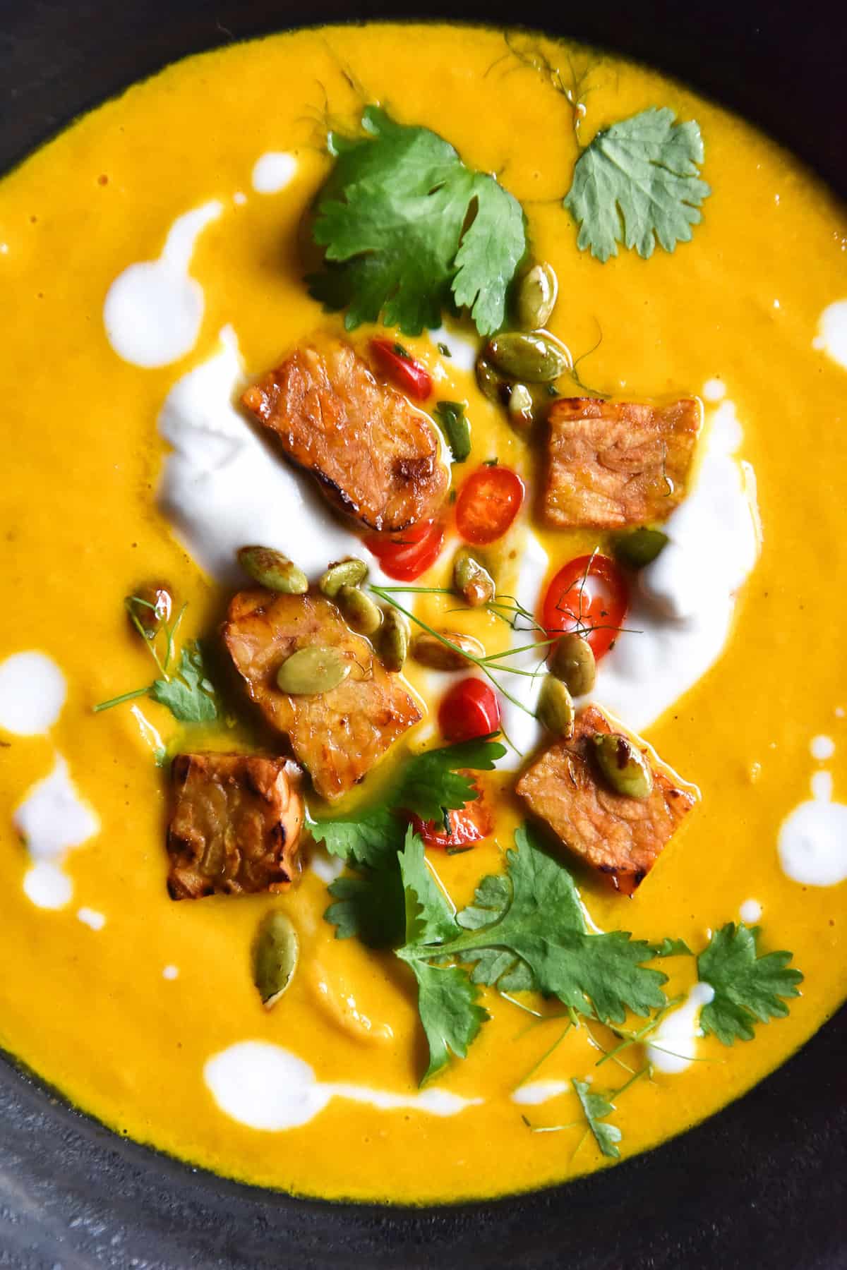 A close up aerial photo of a bowl of vegan Thai carrot soup with crunchy tempeh bits, cream, herbs and chopped chilli.