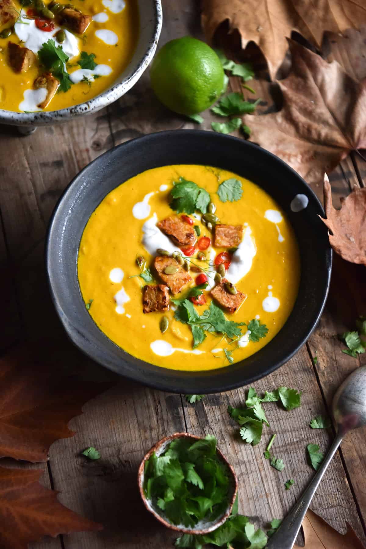 Vegan Thai carrot soup with crunchy tempeh bits. The soup is in a dark ceramic bowl on a wooden table surrounded by extra herbs and fall leaves.