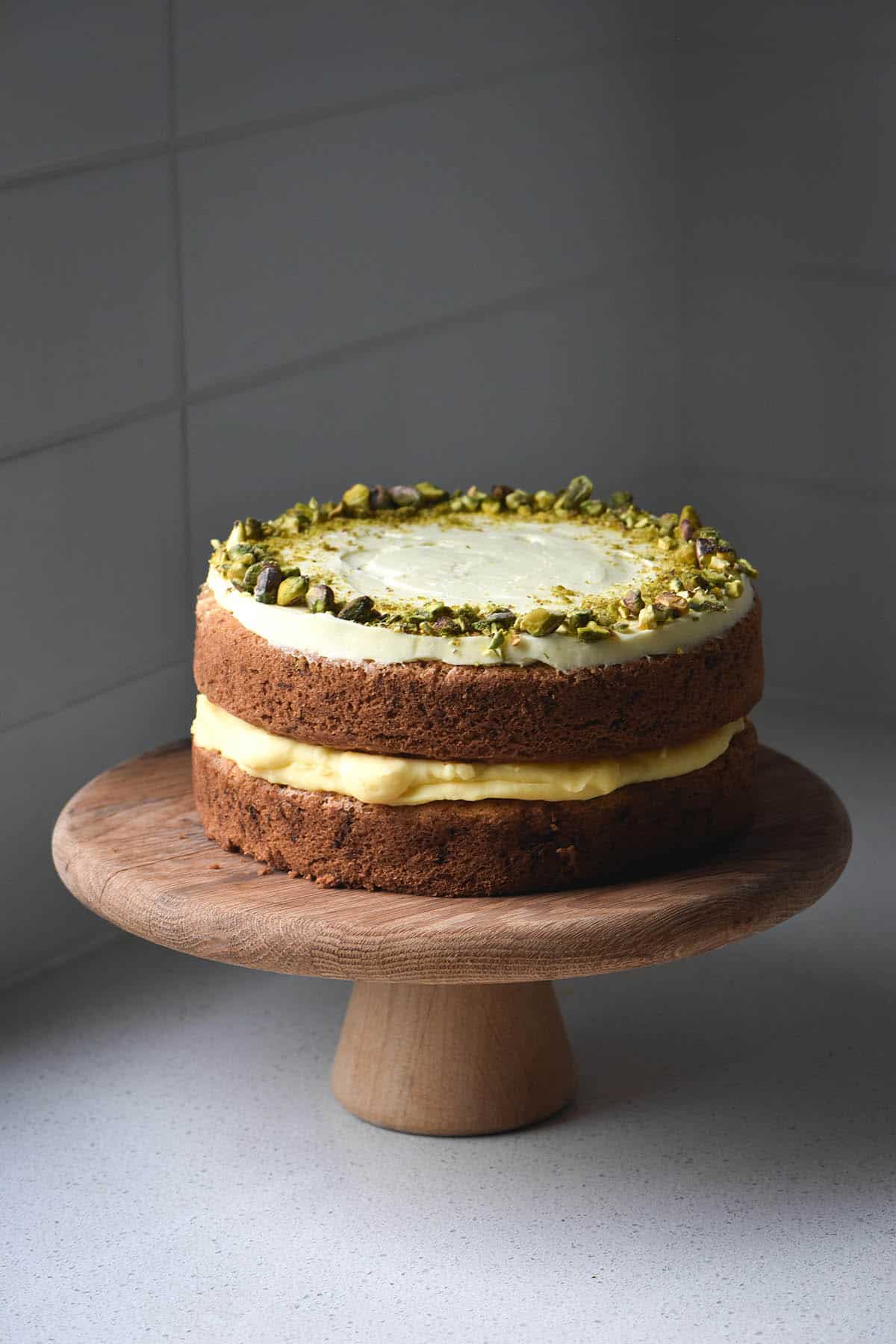 A side on view of a gluten free zucchini cake filled with lime curd and topped with cream cheese icing and pistachios. The cake sits on a wooden cake stand on a white benchtop