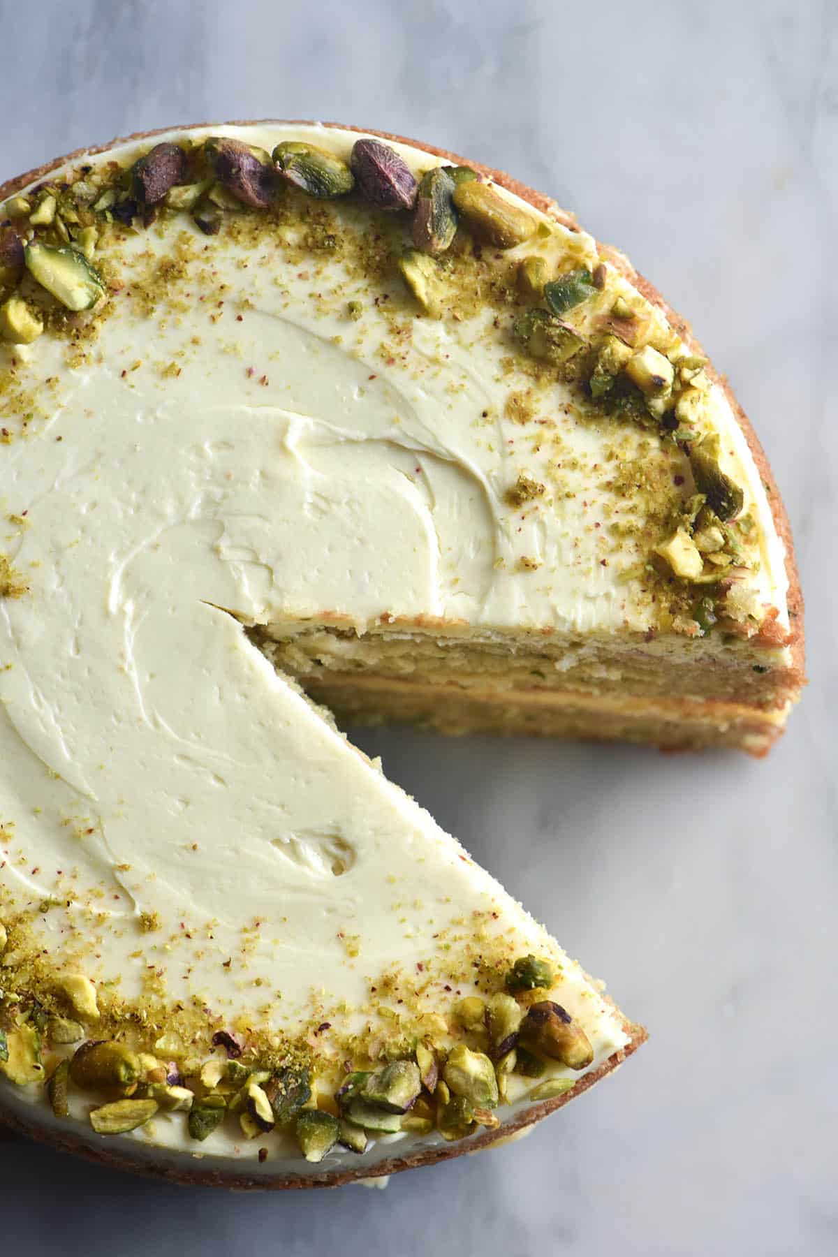 An aerial close up image of a zucchini cake topped with cream cheese icing and a ring of chopped pistachios. The cake sits atop a white marble table and a slice has been removed from the bottom right corner.