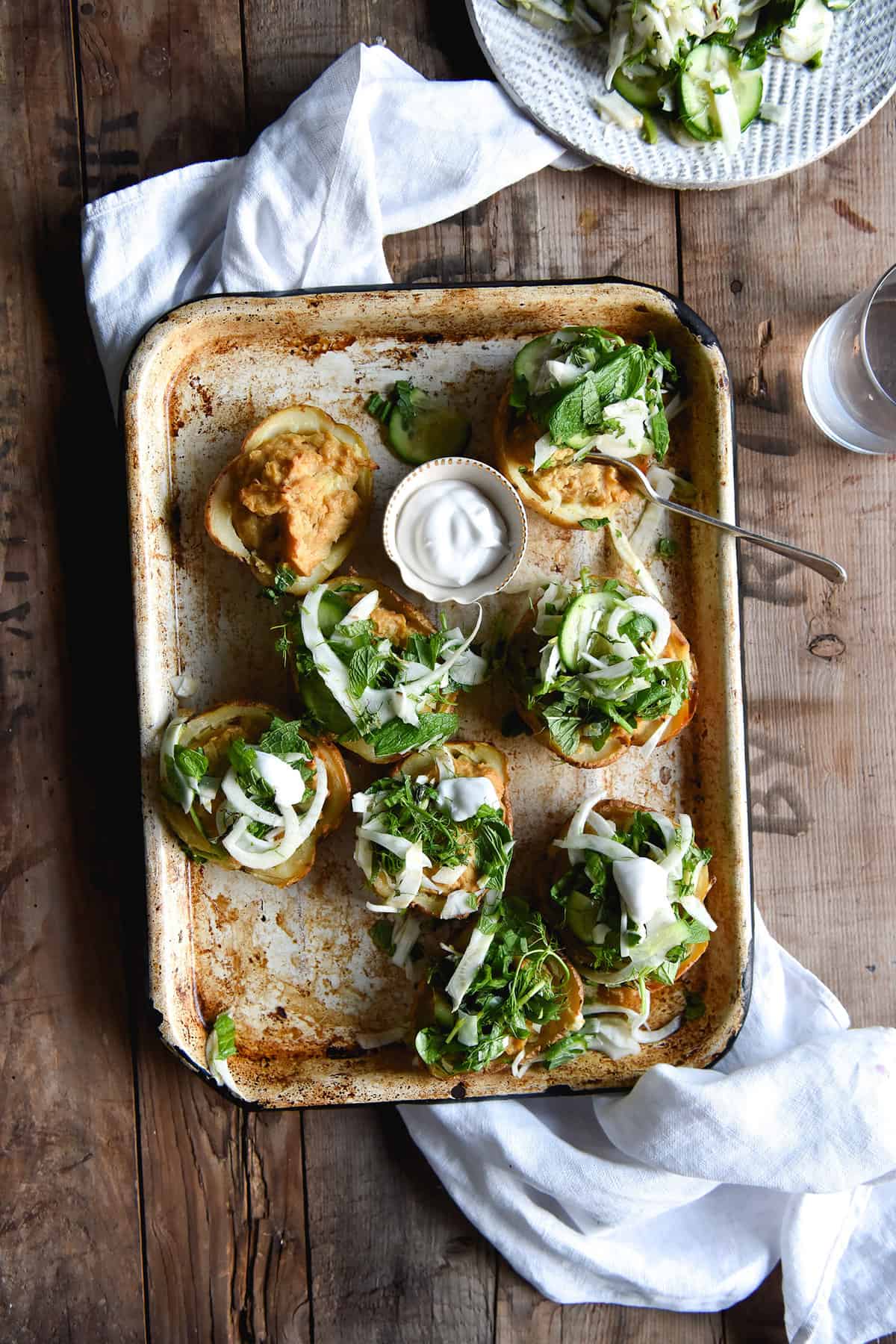 An aerial image of vegan cheesy baked potatoes with a green salsa on a rustic baking tray atop a wooden table and a white linen tea towel.