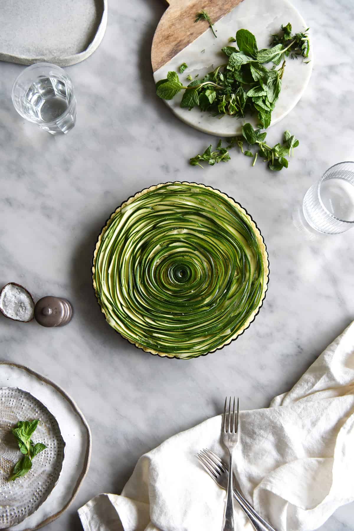 An aerial image of a vegan gluten free zucchini swirl tart on a white marble table. The tart is surrounded by white ceramic plates, water glasses, salt and pepper and a white linen cloth. 
