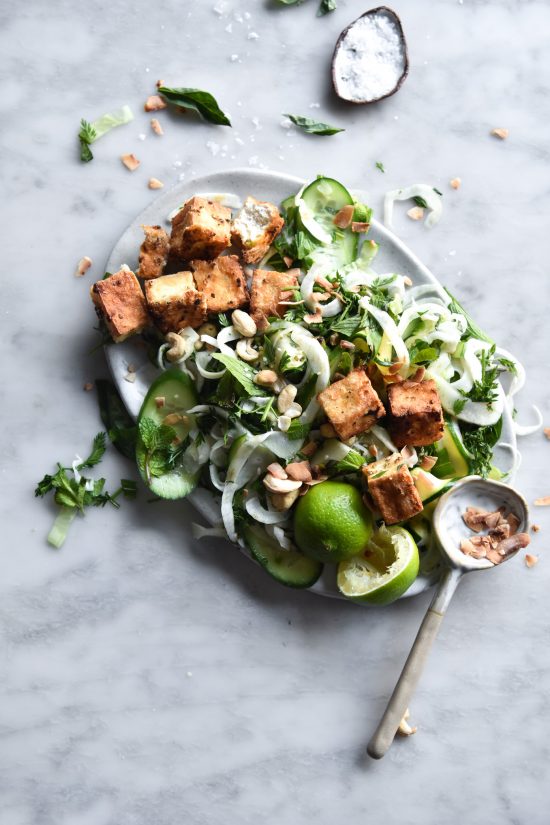 An aerial image of a crispy tofu salad with fresh greens, cucumbers, a ginger satay some fresh peanuts for topping. The salad sits on a white marble table.