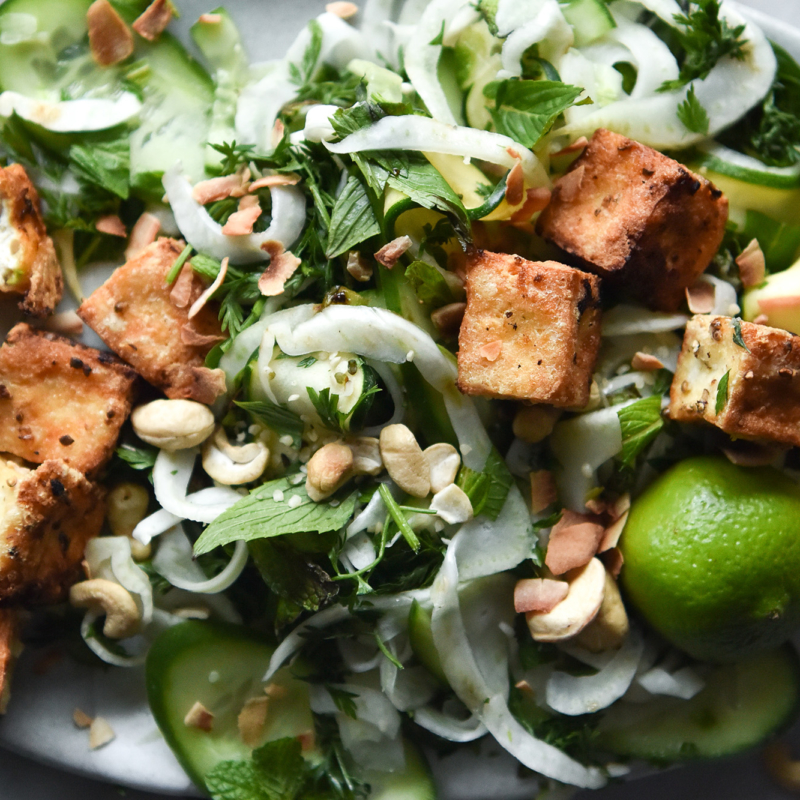 A close up photo of a green salad topped with crispy nuggets of pan fried tofu. The salad sits on a white plate atop a white marble table