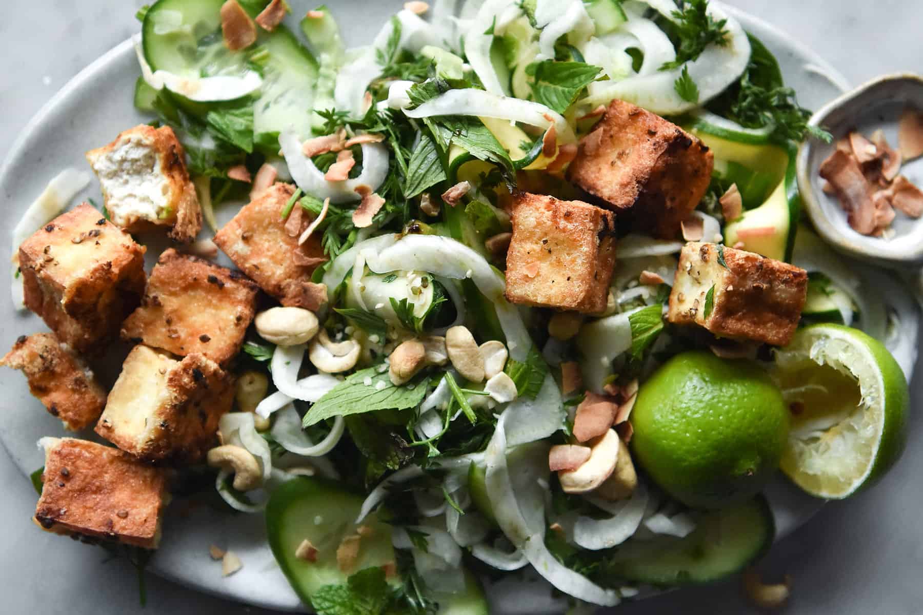 A close up photo of a green salad topped with crispy nuggets of pan fried tofu. The salad sits on a white plate atop a white marble table