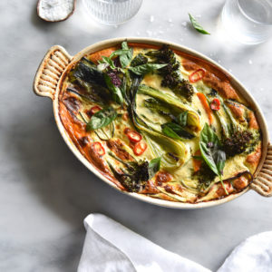 A green Thai frittata baked in an oval baking dish sits on a white marble table surrounded by water glasses and a white linen tablecloth