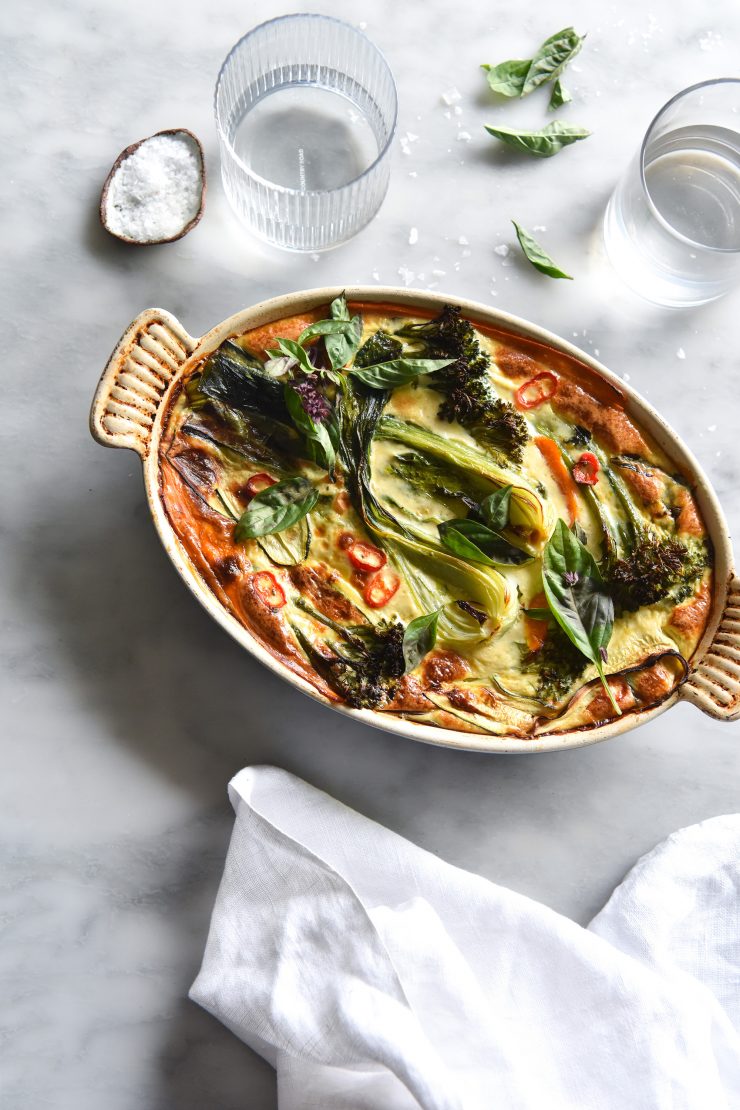 A green Thai frittata baked in an oval baking dish sits on a white marble table surrounded by water glasses and a white linen tablecloth