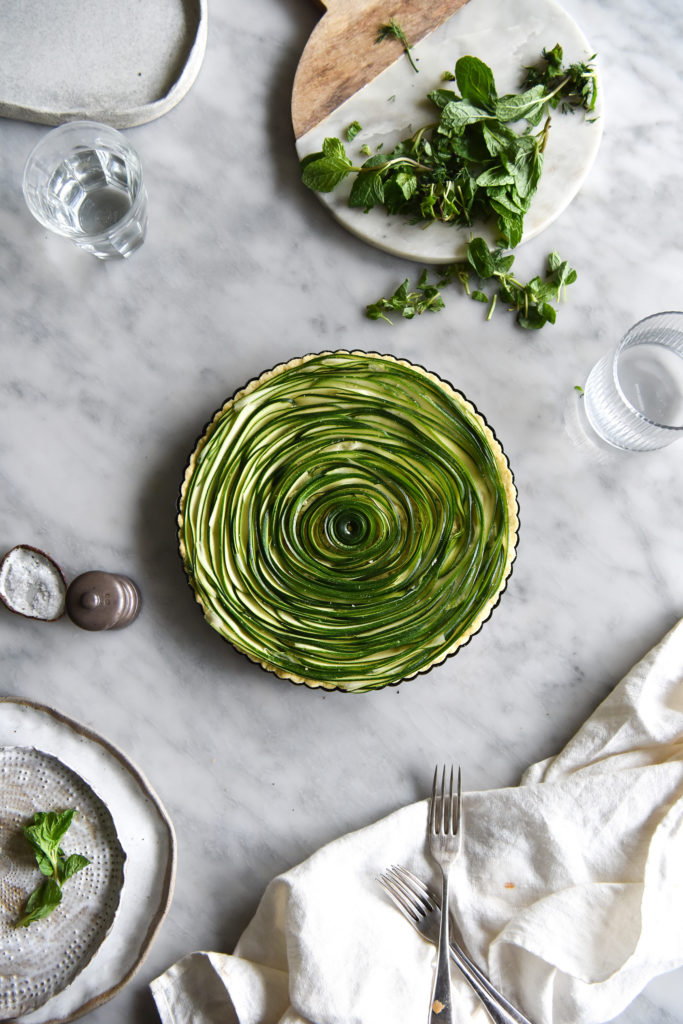 Grain free zucchini swirl tart, surrounded by plates and place on a light marble backdrop