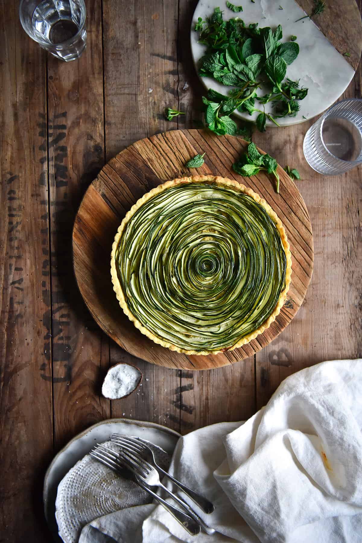 An aerial image of a zucchini swirl tart on a wooden board atop a wooden table. The tart is surrounded by glasses of water, extra mint on a marble board, plates and a white linen cloth.