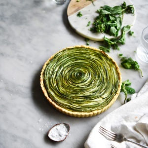An aerial view of a gluten free zucchini swirl tart on a white marble table. The tart is surrounded by white plates, linen and extra chopped mint.