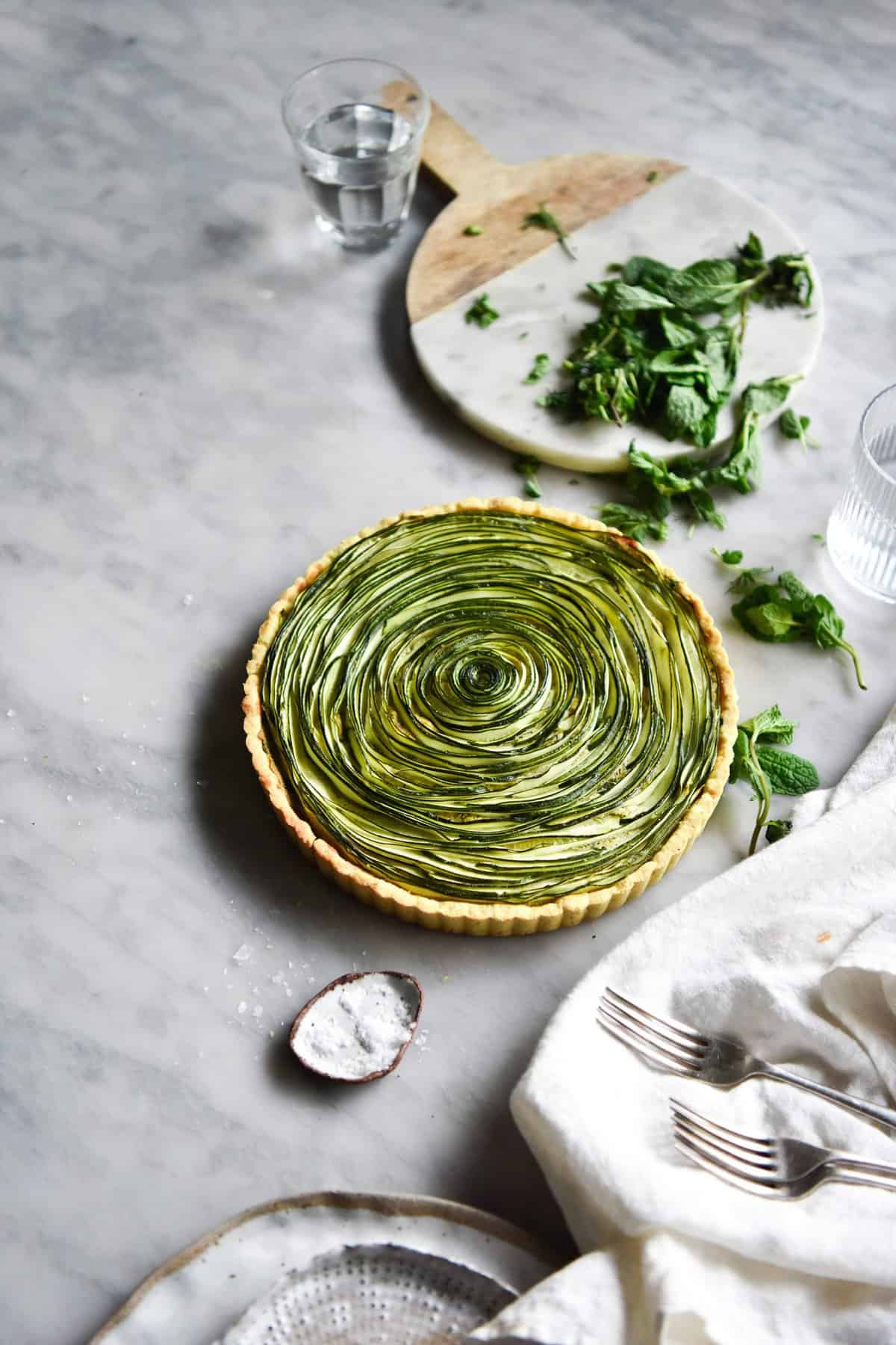 An aerial view of a gluten free zucchini swirl tart on a white marble table. The tart is surrounded by white plates, linen and extra chopped mint.