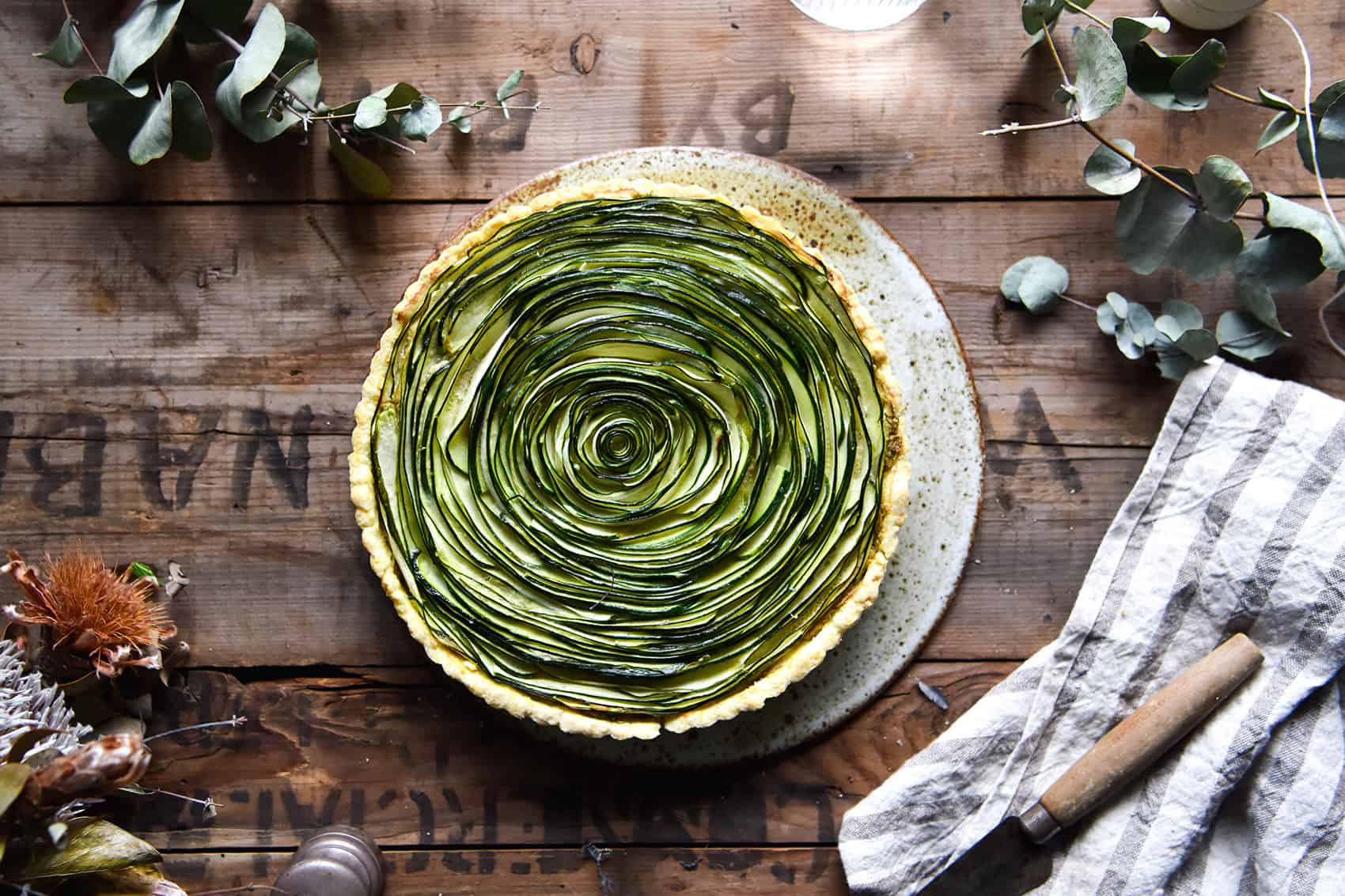 Grain free zucchini swirl tart on a wooden, moody backdrop surrounded by fall foliage and a linen tea towel