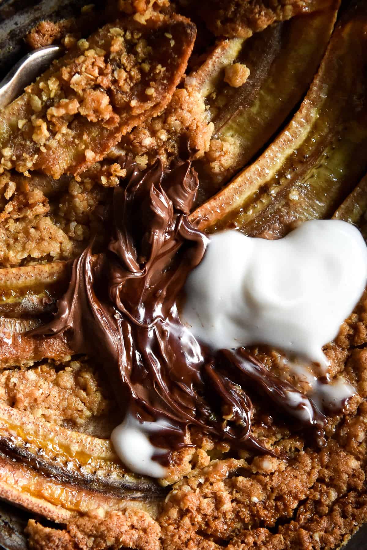 An aerial macro image of a gluten free breakfast crumble on bananas. The crumble is topped with Nutella and yoghurt.