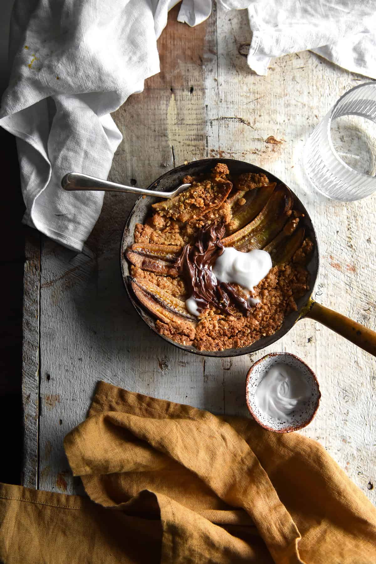 An aerial image of a gluten free breakfast crumble on bananas. The crumble is topped with Nutella and yoghurt.The skillet sits on a light coloured wooden table. 