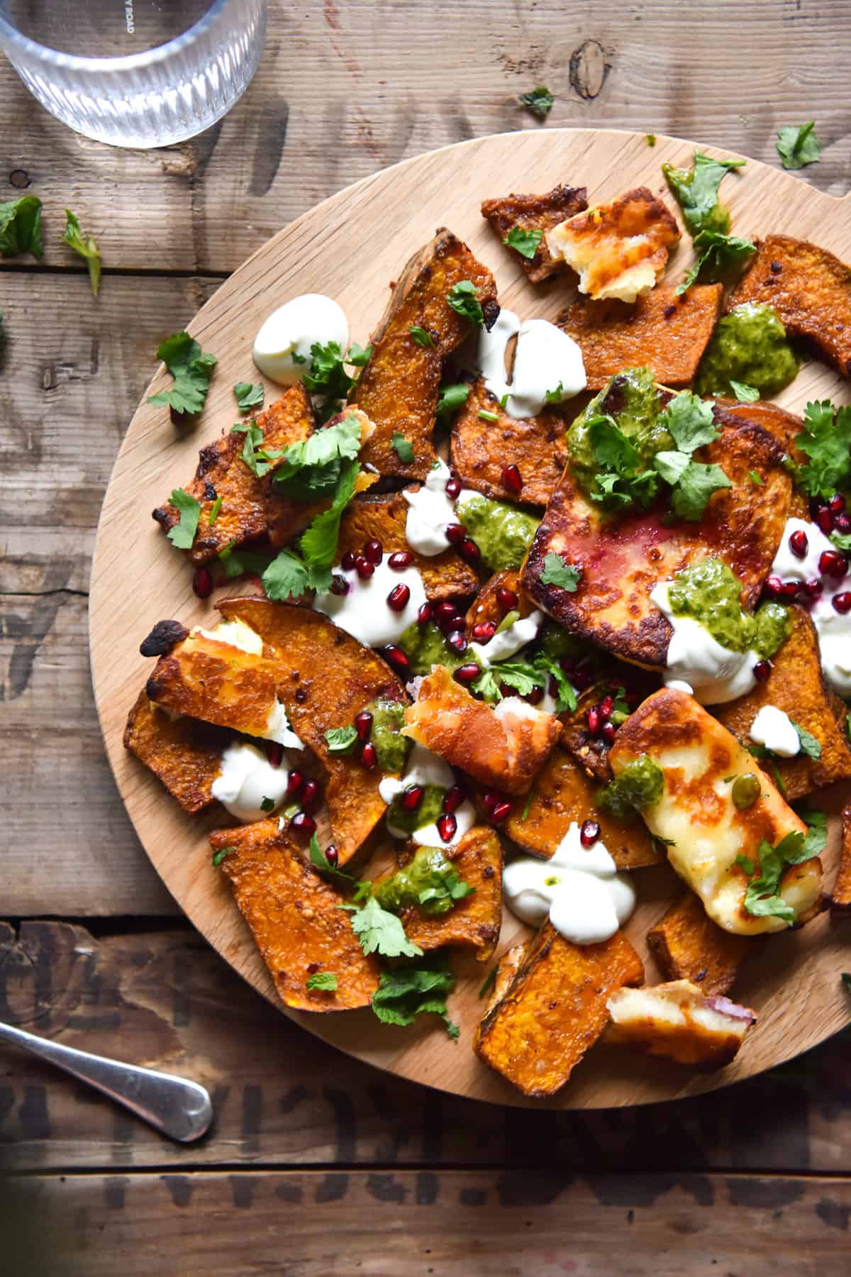 An aerial photo of a spiced pumpkin, halloumi, pomegranate and herb sauce salad on a wooden board against a wooden backdrop. A glass of water sits to the top left of the image.