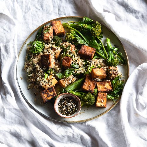 An aerial view of a plate of miso glazed tofu, broccolini and furikake rice bowls on a white linen tablecloth
