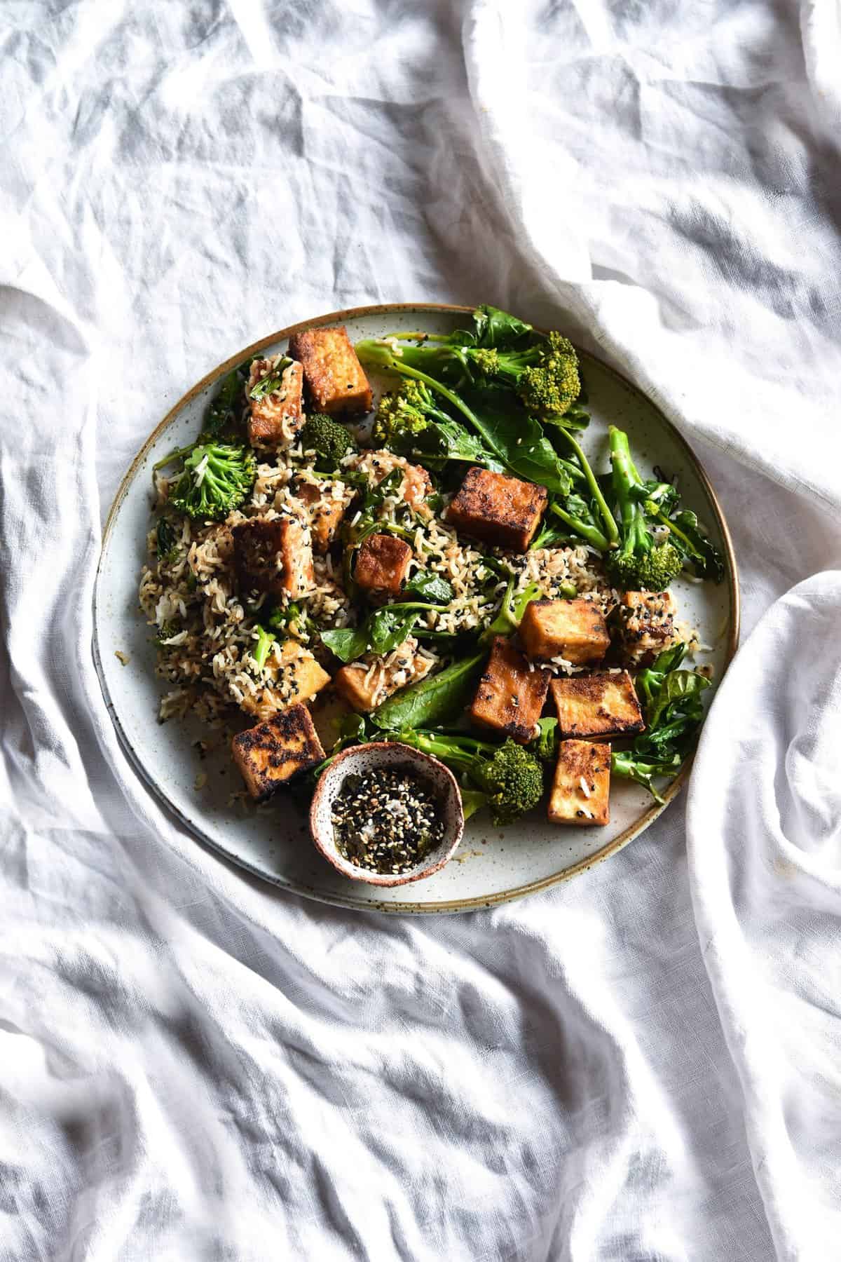 An aerial view of a plate of miso glazed tofu, broccolini and furikake rice bowls on a white linen tablecloth