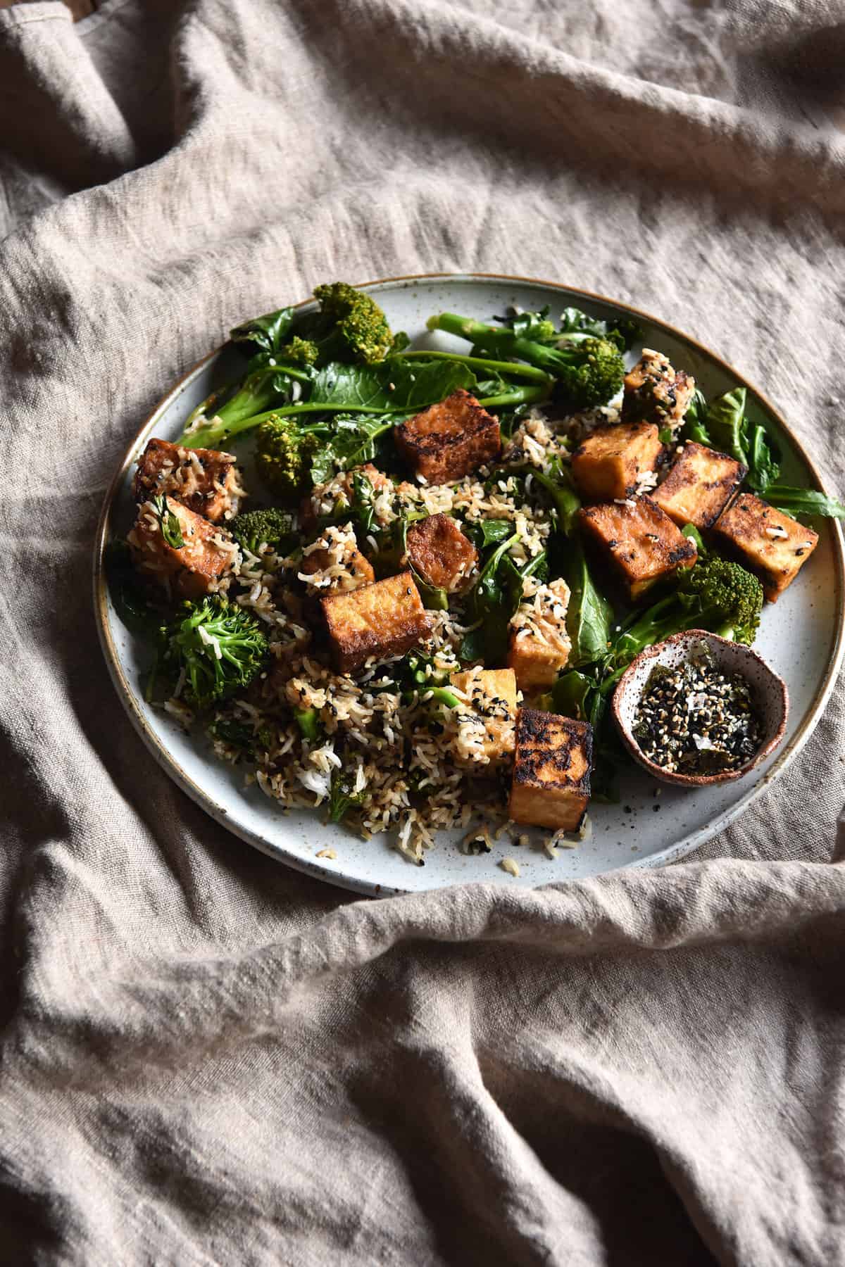 Furikake rice with miso glazed tofu and broccolini on a white ceramic plate that sits on a textured bone coloured linen tablecloth.