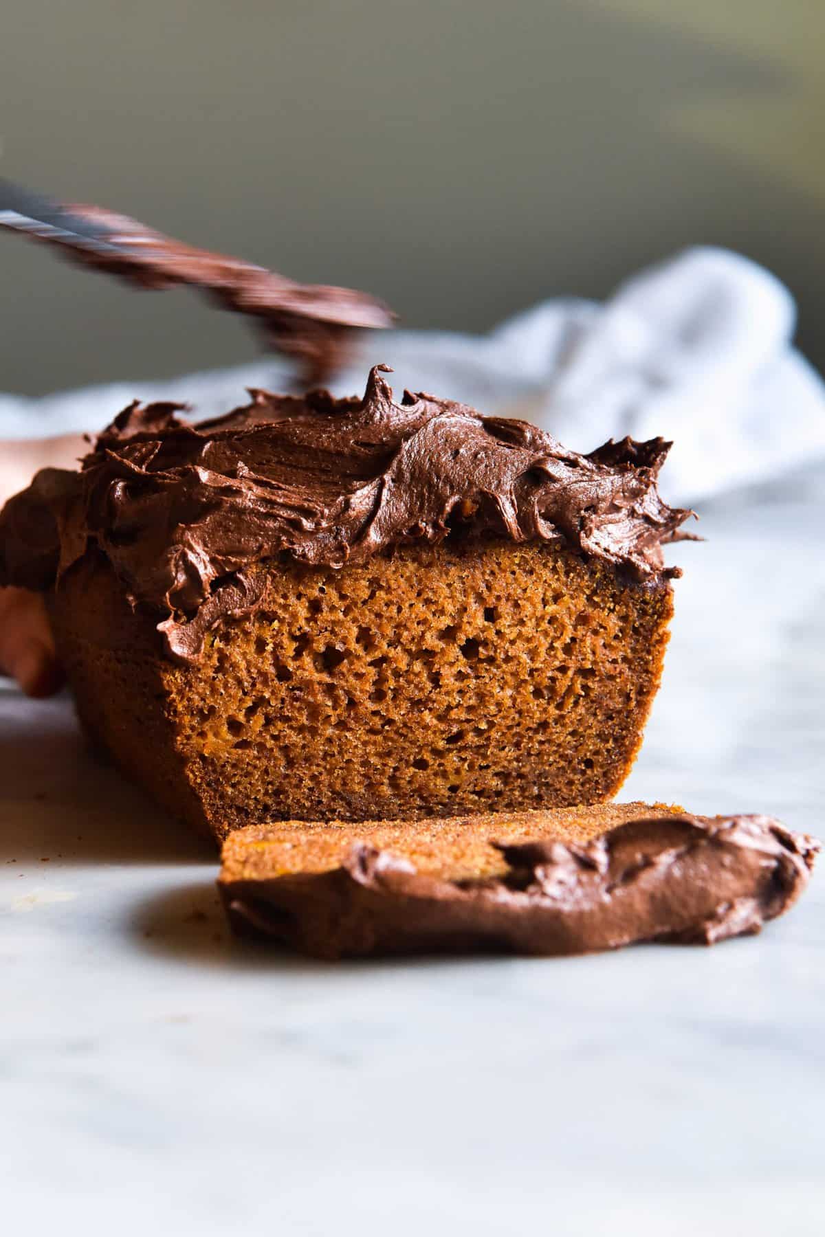 A side on view of a gluten free pumpkin loaf topped with chocolate chai icing. The loaf has been sliced to reveal the bright orange crumb. It sits on a white marble table against a light backdrop.