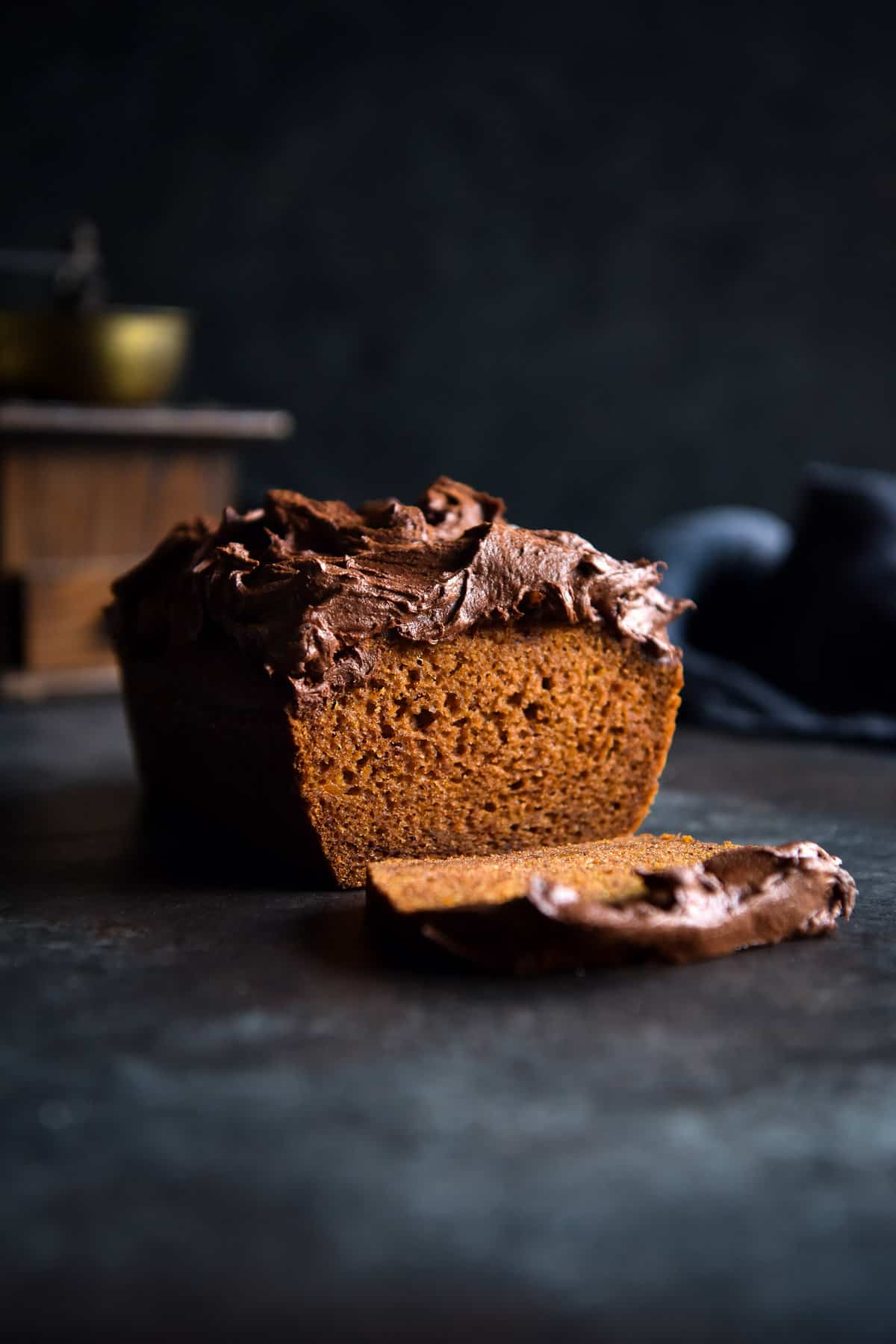 A side on view of a gluten free pumpkin loaf topped with chocolate chai icing. The loaf has been sliced to reveal the bright orange crumb. It sits on a dark blue table against a dark blue backdrop.